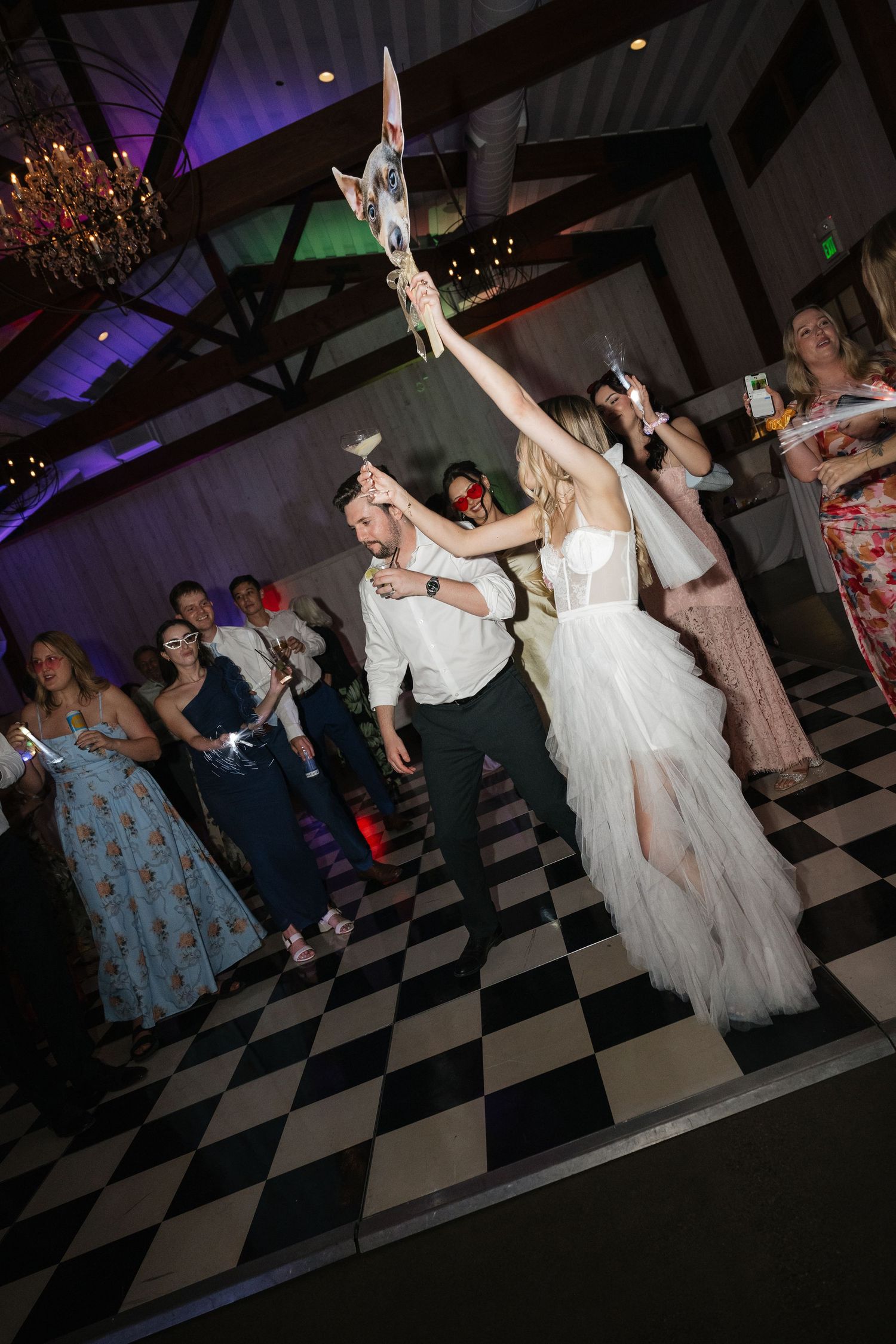 Wedding guests dance together on a black and white checkered dance floor under purple lighting at a reception celebration.
