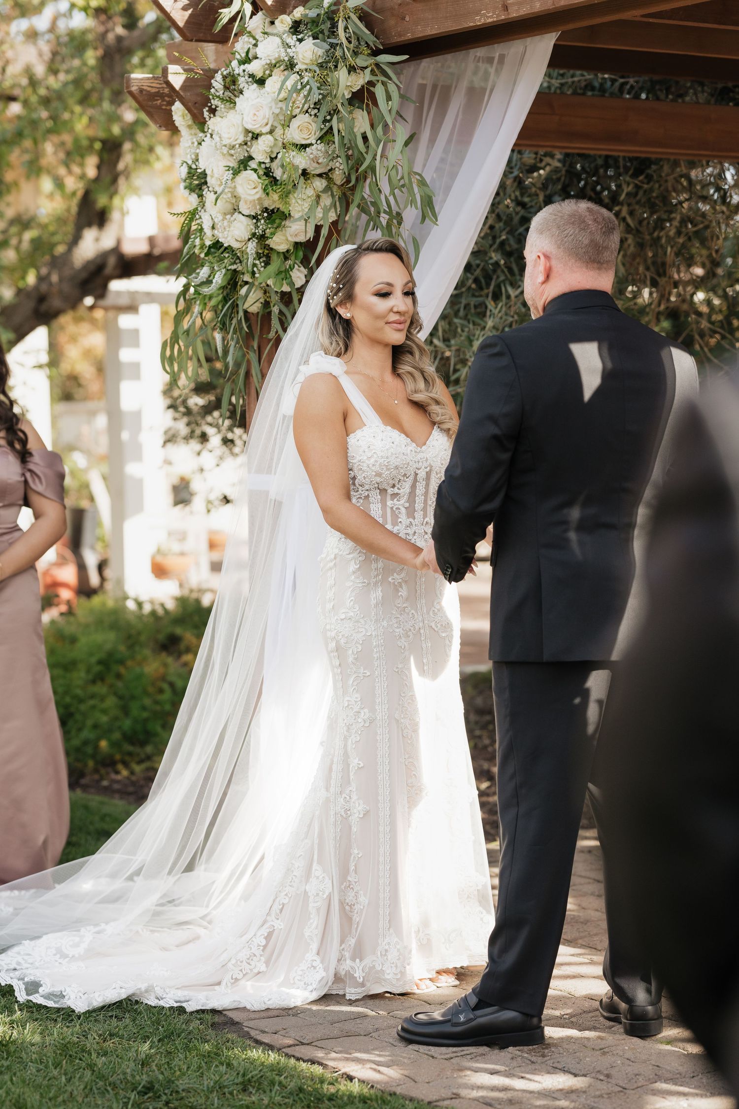Wedding ceremony under wooden pergola decorated with greenery and white flowers.