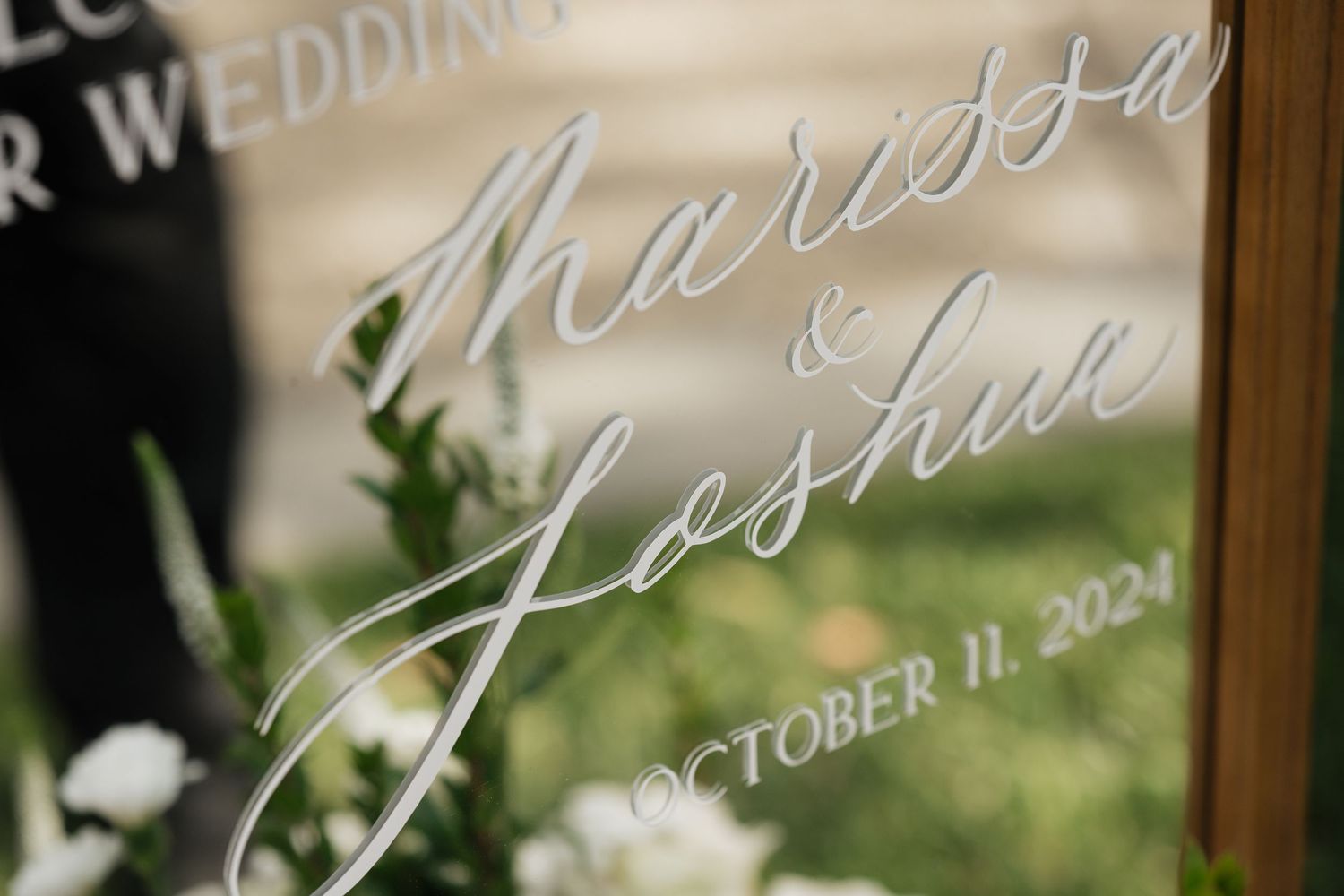 Elegant script lettering on clear acrylic wedding sign with natural background.