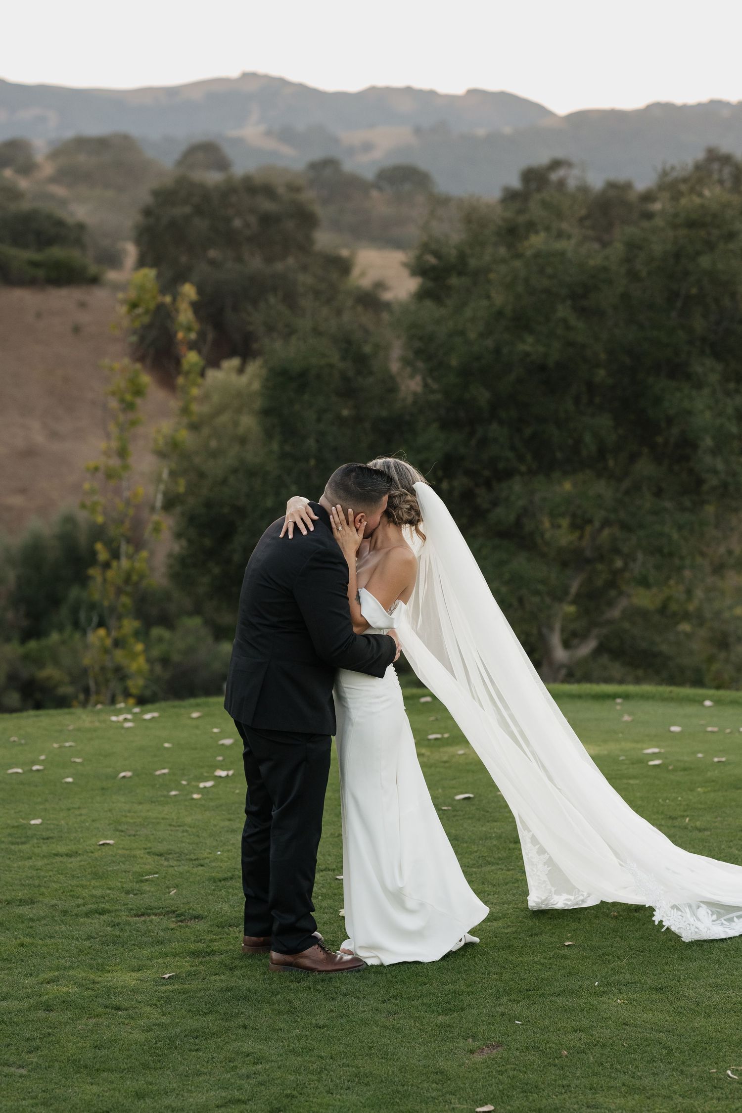 A couple in wedding attire shares an intimate moment on a grassy hillside with mountains in the background.