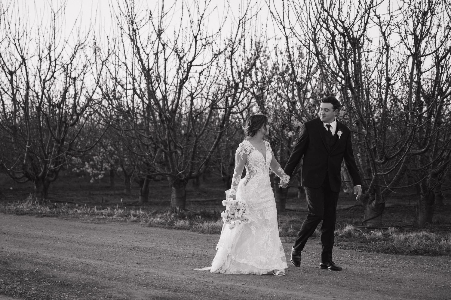 Black and white photograph of wedding couple walking through bare winter orchard.