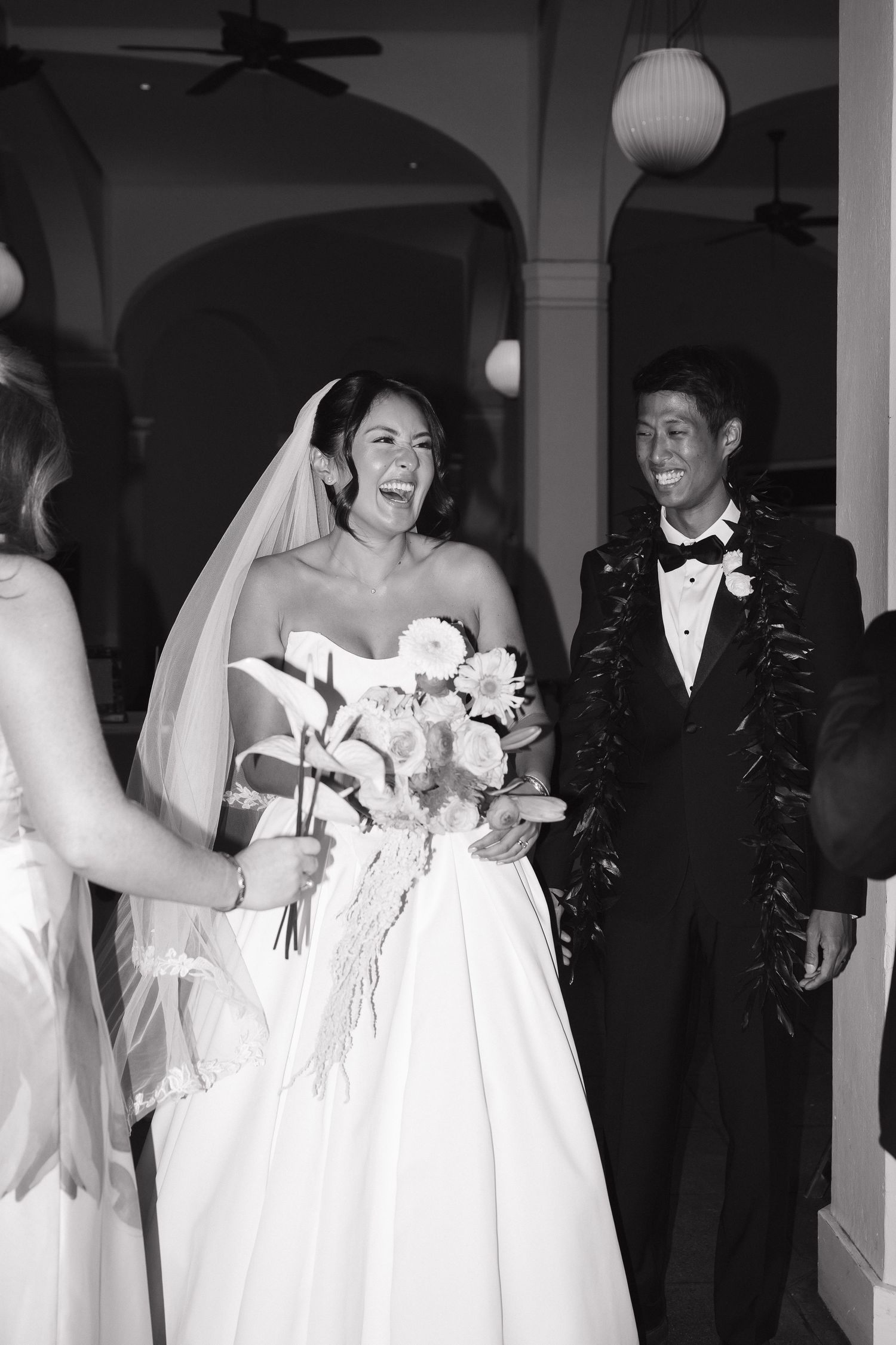 Black and white photo of a joyful bride in a white gown and groom in a tuxedo with lei, laughing after their wedding ceremony.