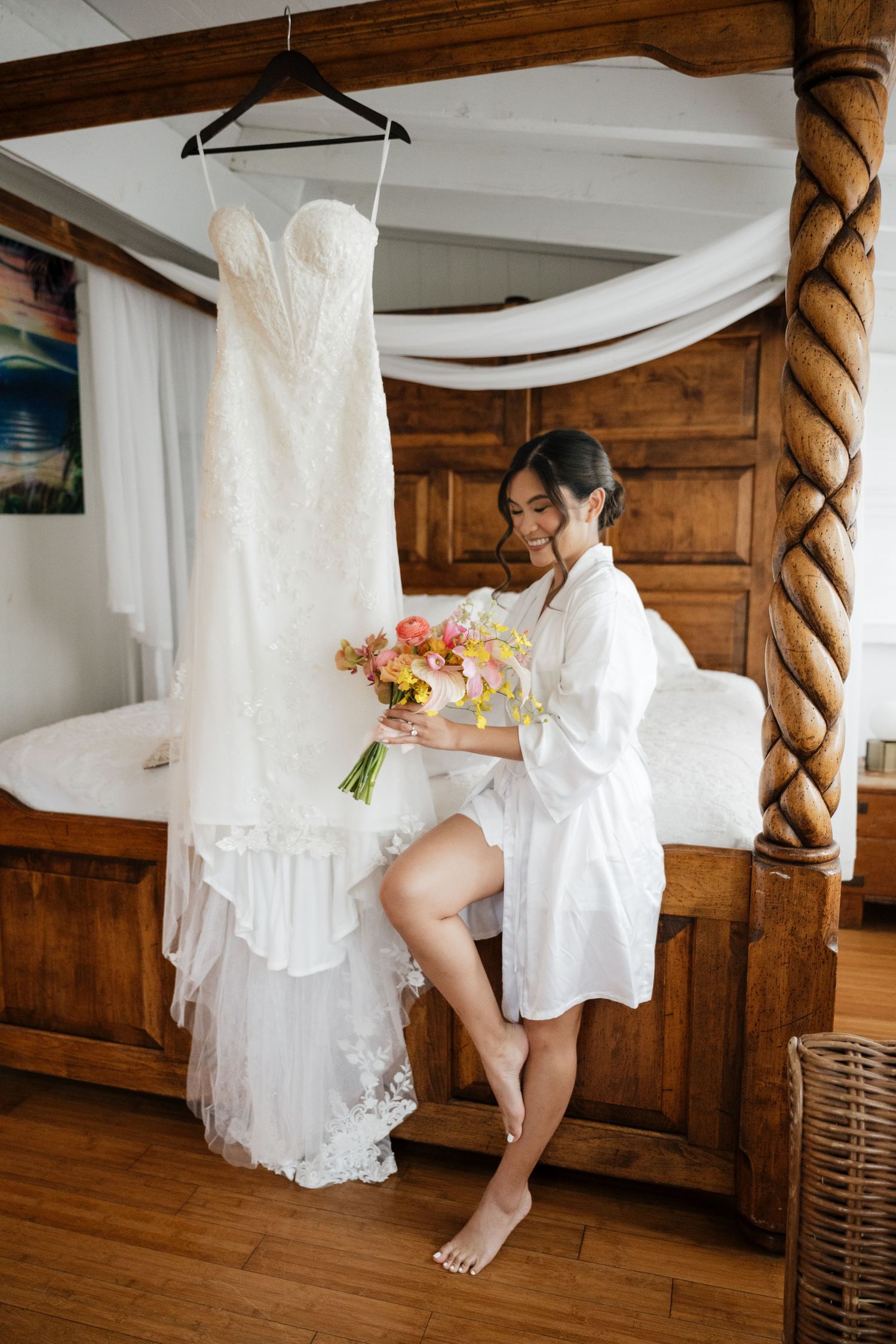 Bride in white robe holds colorful bouquet beside her wedding dress hanging on a four-poster bed.