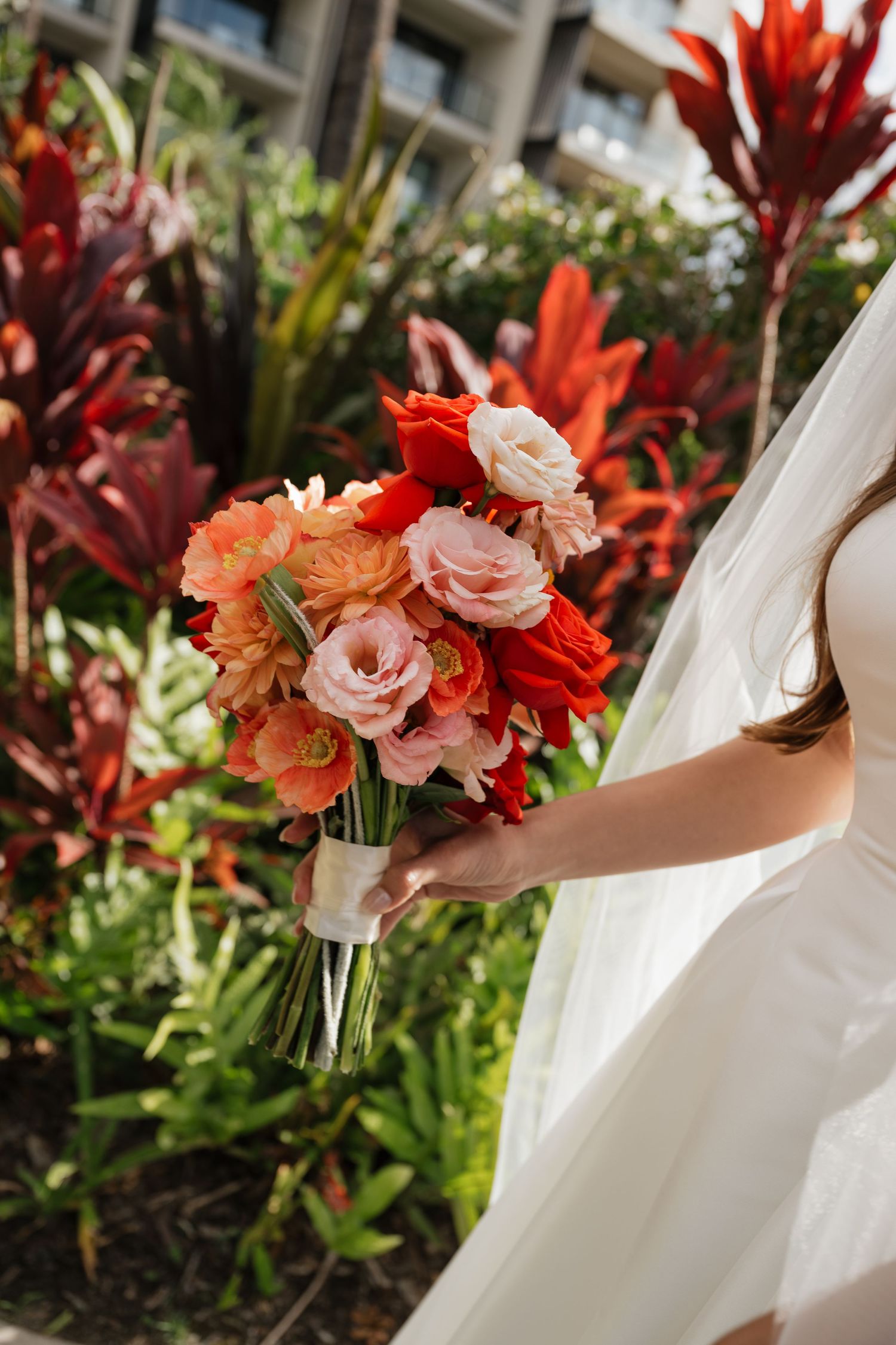 Vibrant red and coral colored wedding bouquet with roses and tropical flowers against lush garden backdrop.