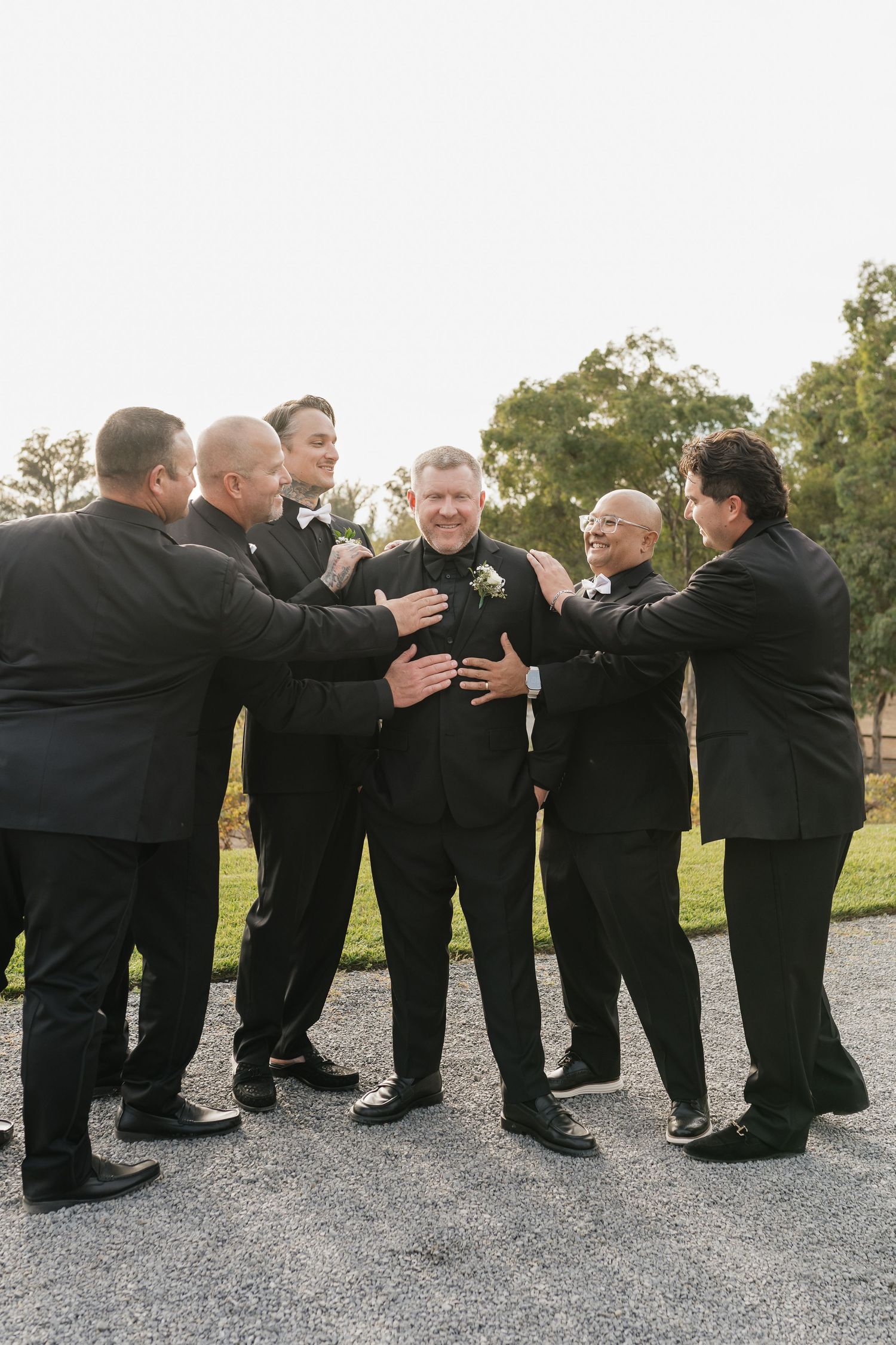 Groomsmen in black tuxedos share a fun group hug moment outdoors.