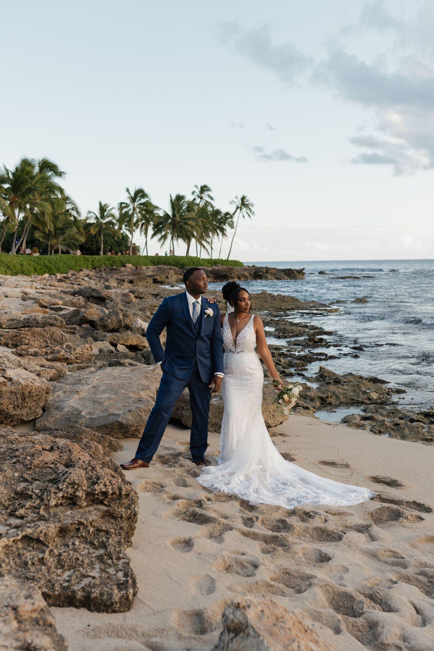 A romantic wedding photo on a rocky beach with palm trees and ocean views.