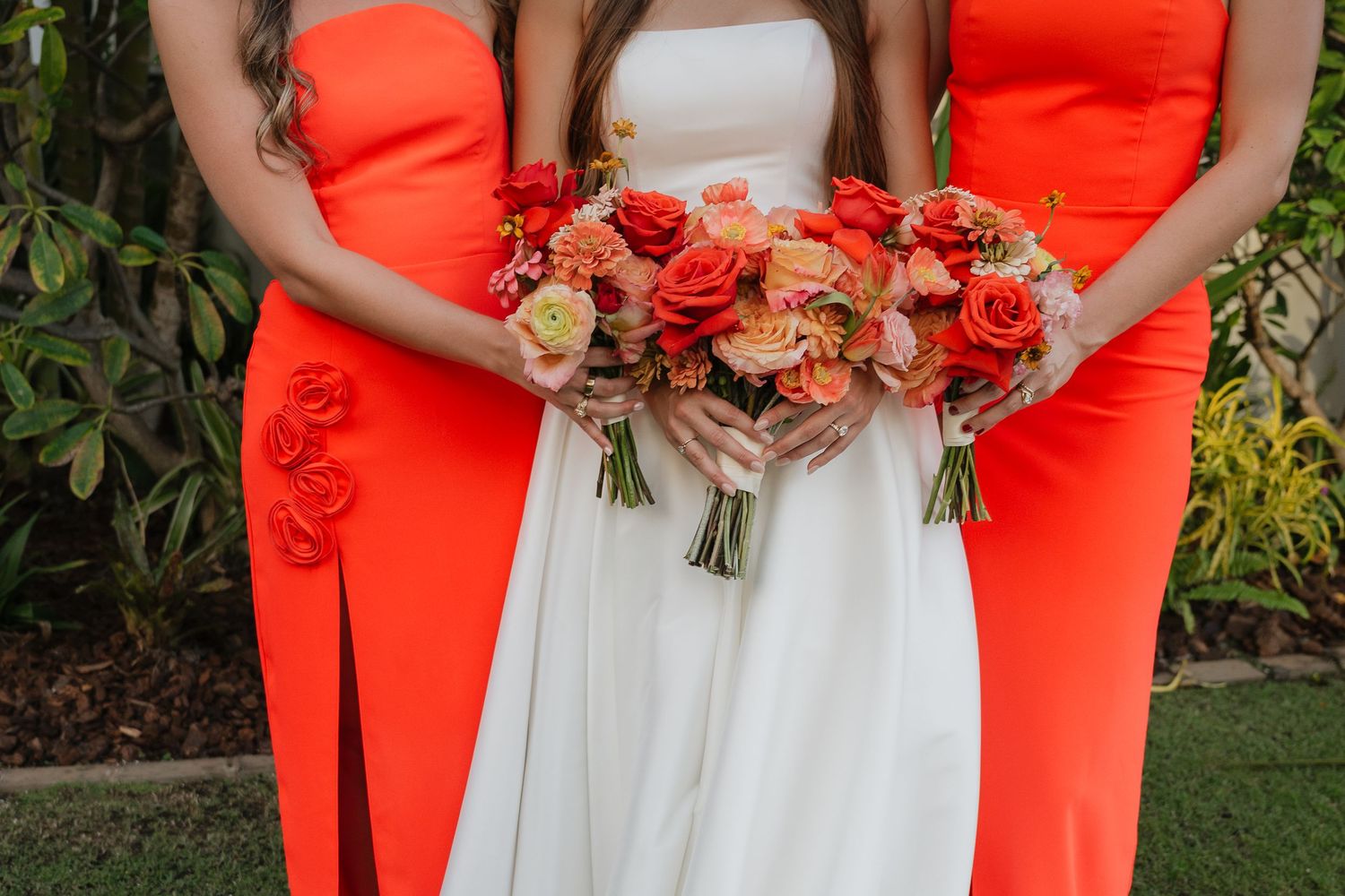 Three bouquets of coral and peach roses held by bridesmaids wearing orange and white dresses at an outdoor wedding.