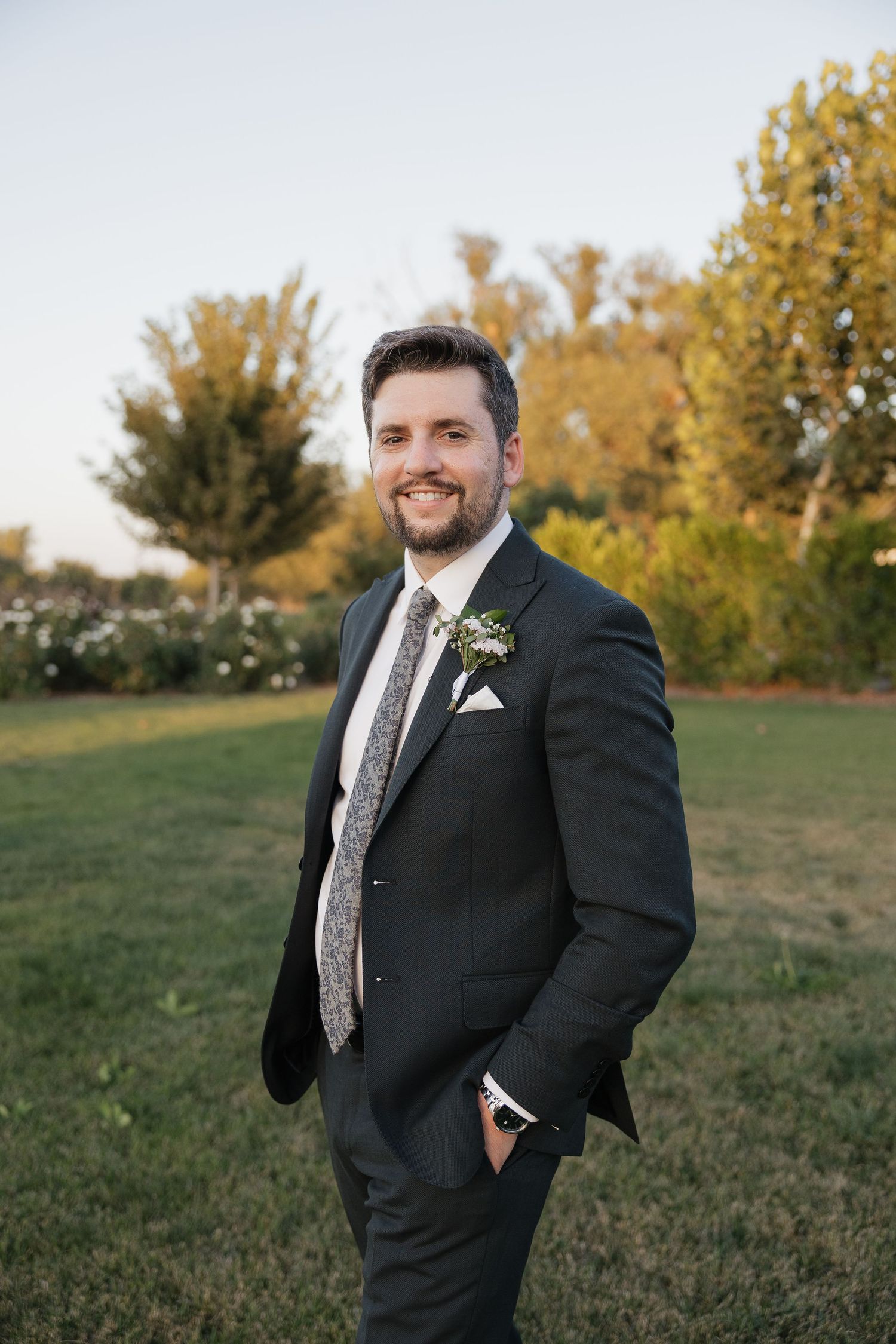 A groom in a dark suit and patterned tie poses outdoors with autumn foliage in the background.