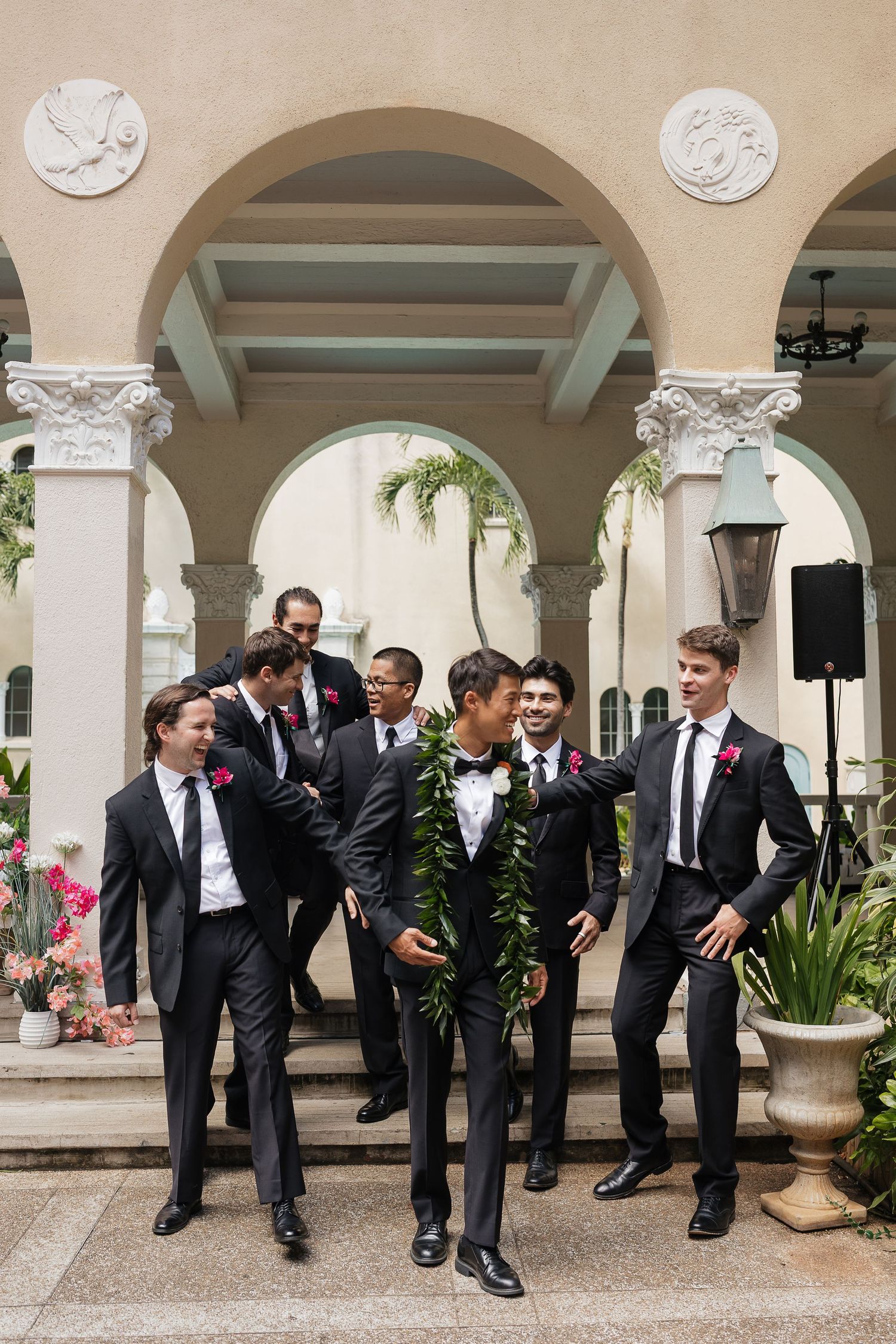 Groom wearing lei and bow tie laughs with groomsmen in black suits with pink boutonnieres outside elegant venue.