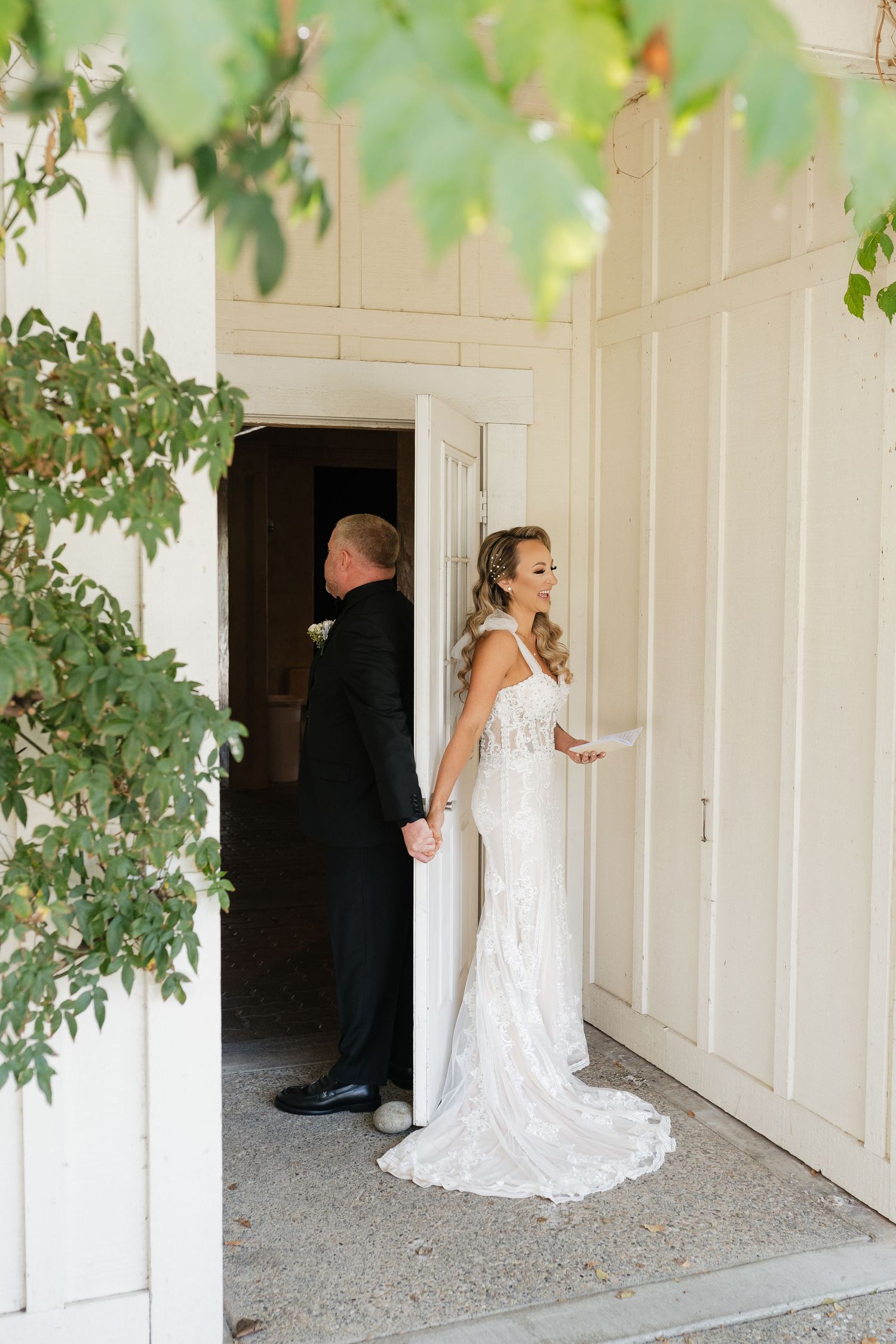 White wedding dress captured from behind against cream-colored wall with foliage overhead.
