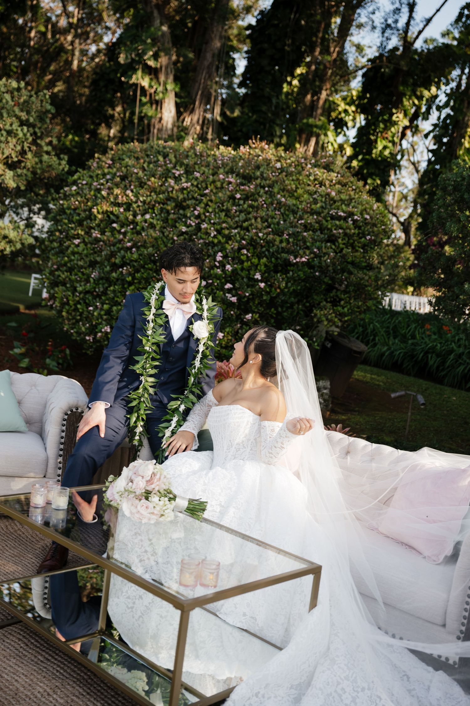 Bride and groom share intimate moment on elegant outdoor lounge seating surrounded by lush greenery.