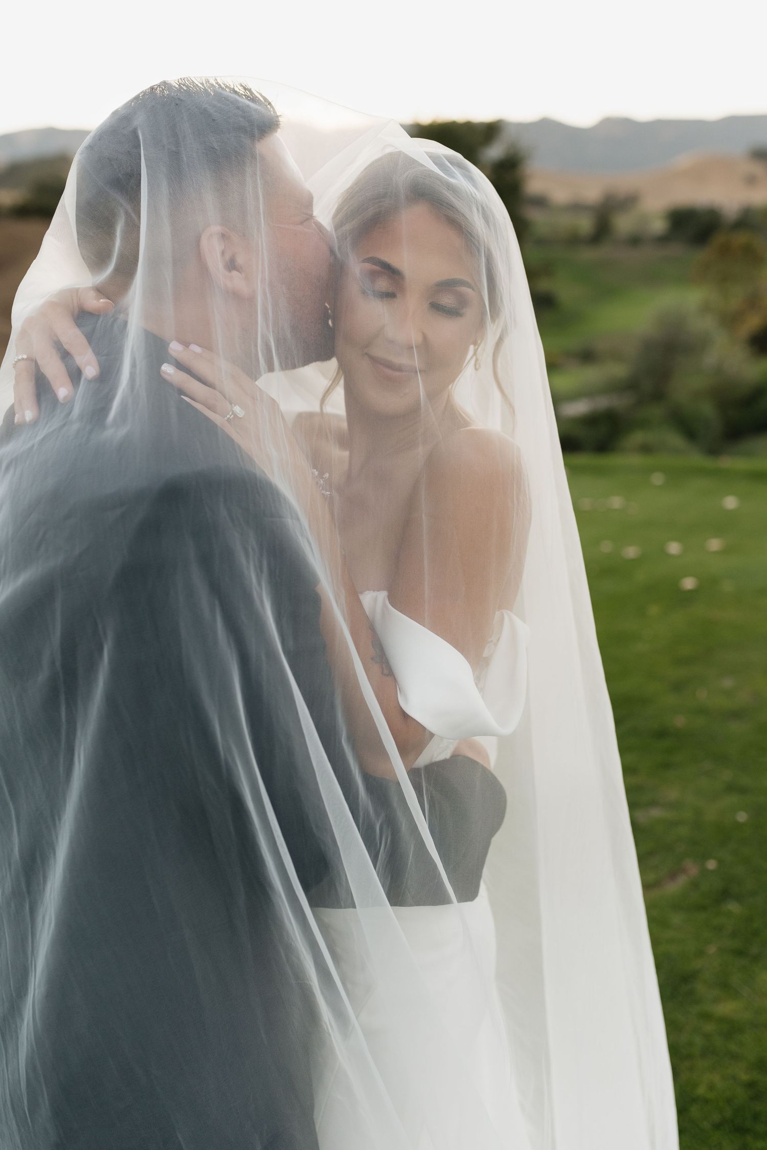 A romantic moment captured through a flowing white veil against a green hillside background.