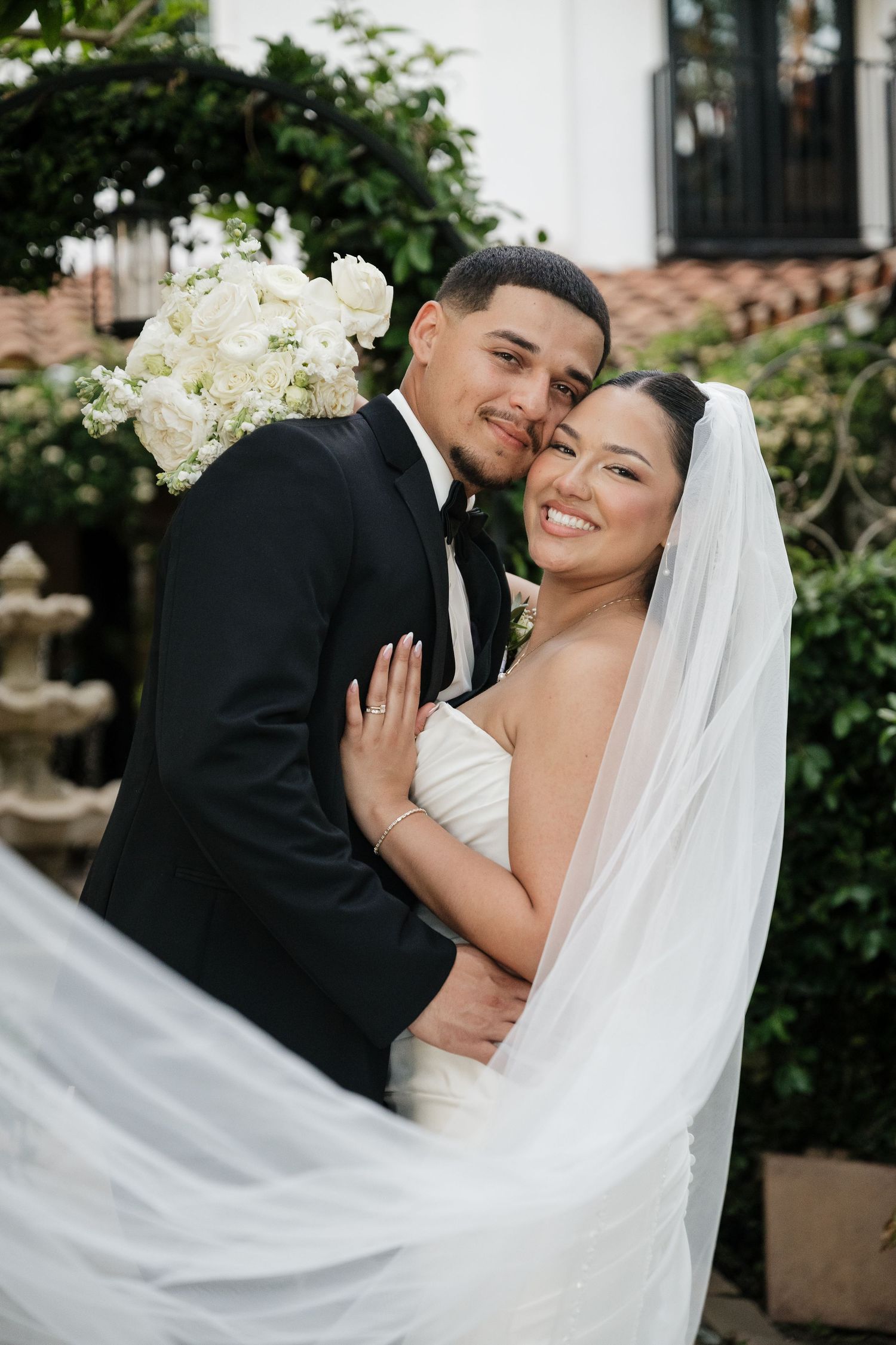 Bride and groom embrace outdoors, she wears a strapless gown with veil, he wears a black tuxedo, with white roses.