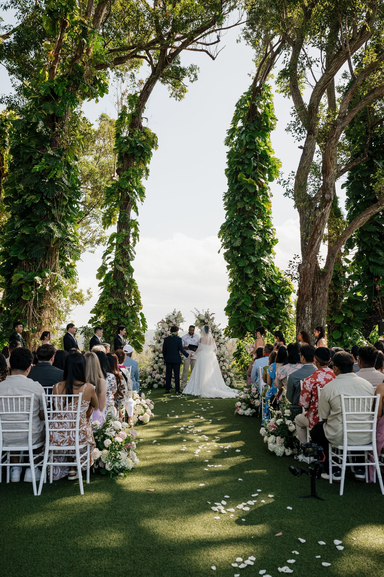 Romantic outdoor wedding ceremony under lush green tree arches with white floral aisle and seated guests.