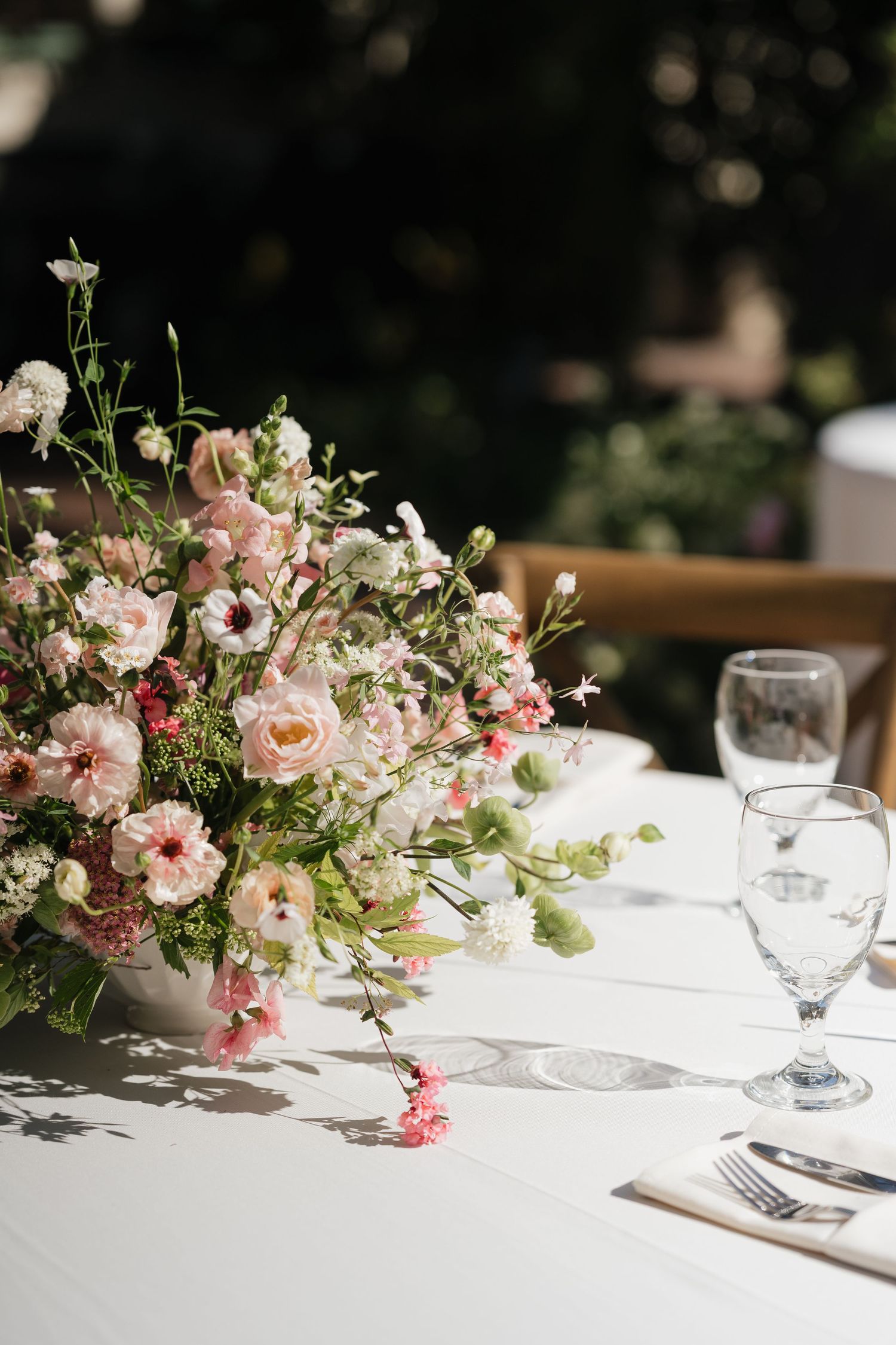 Elegant wedding table centerpiece with pink and white flowers in a white vase, set on a white tablecloth outdoors.