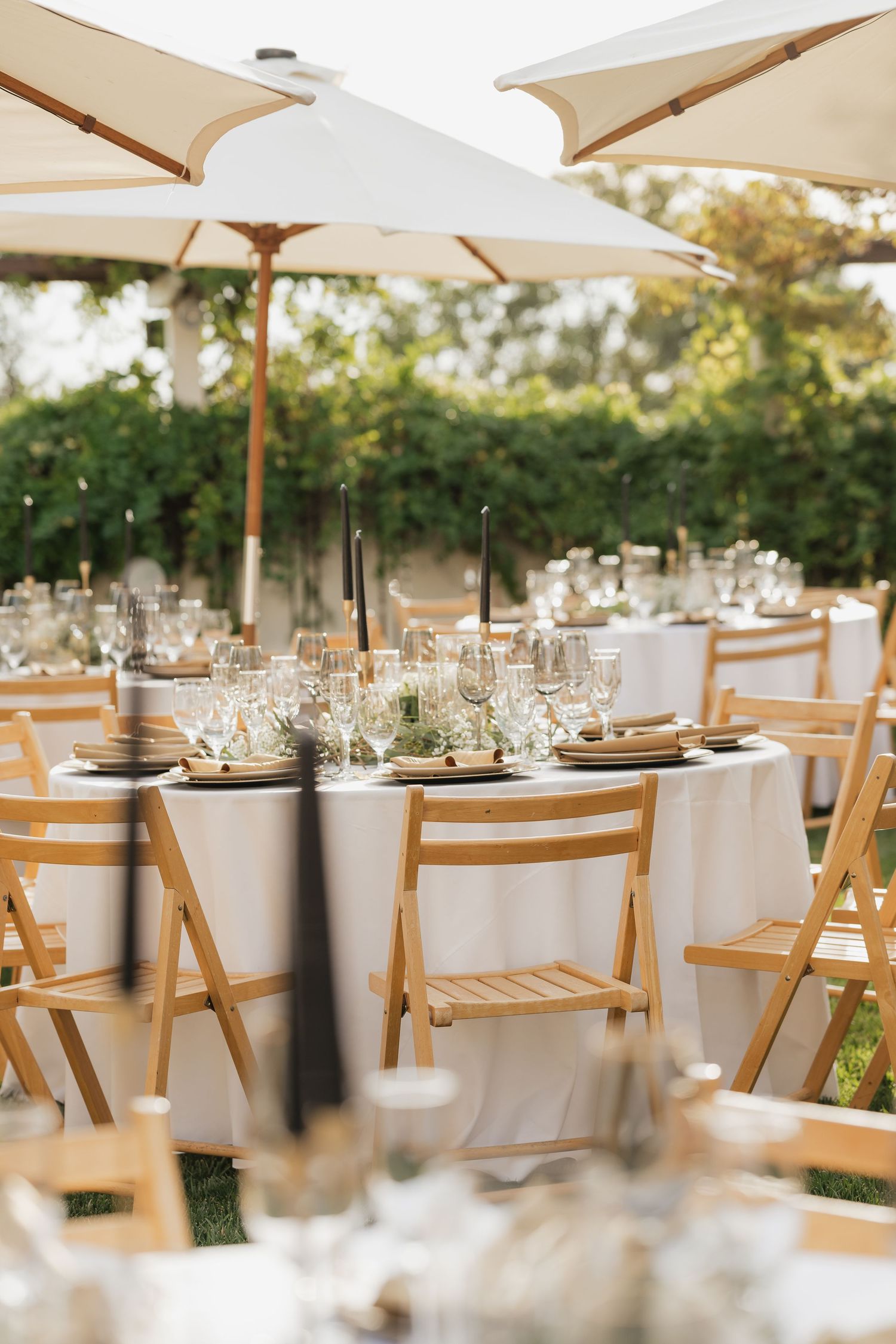 Elegant outdoor reception table settings with wooden chairs and white umbrellas in garden setting.