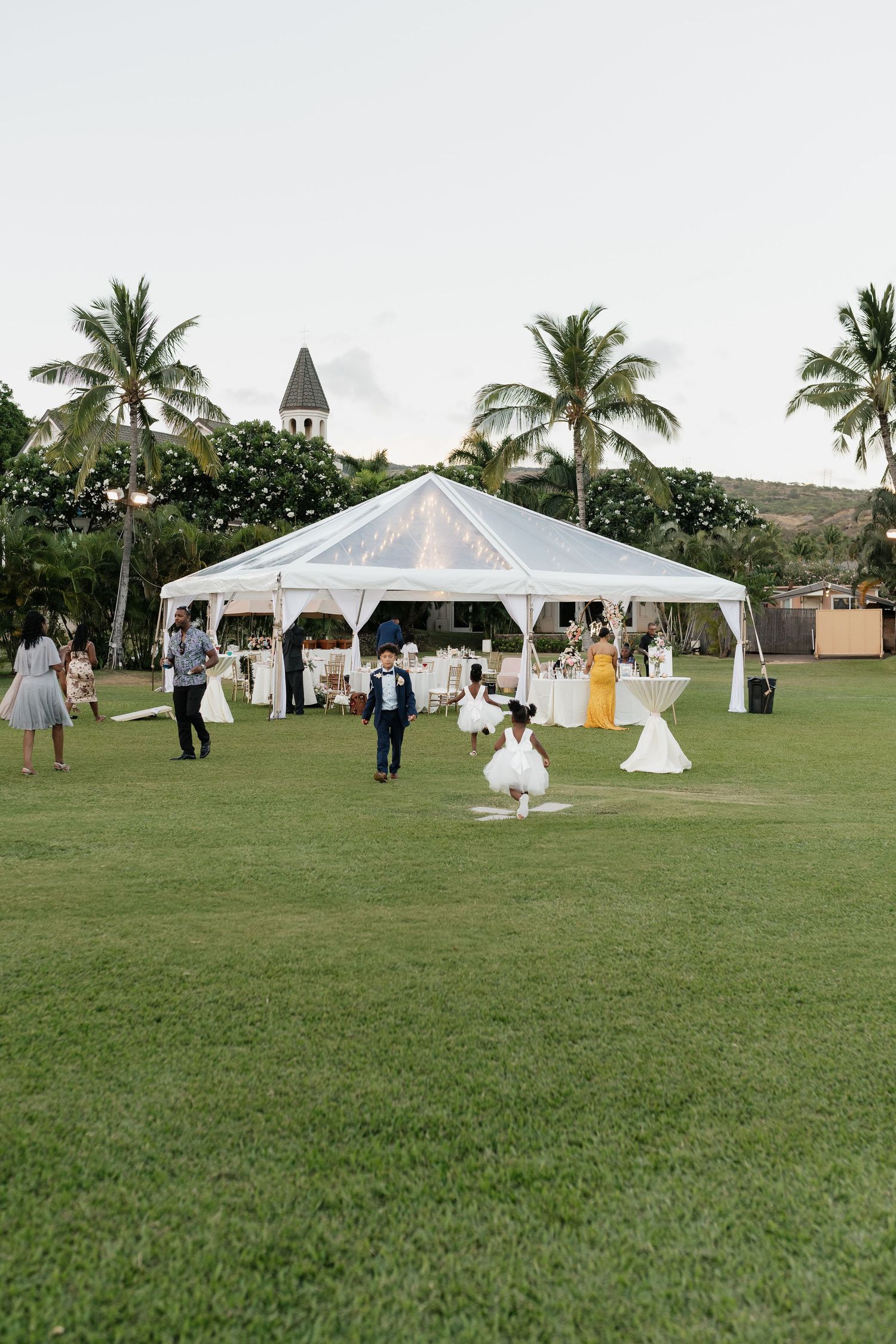 White wedding reception tent set up on a lush green lawn with palm trees in the tropical background.