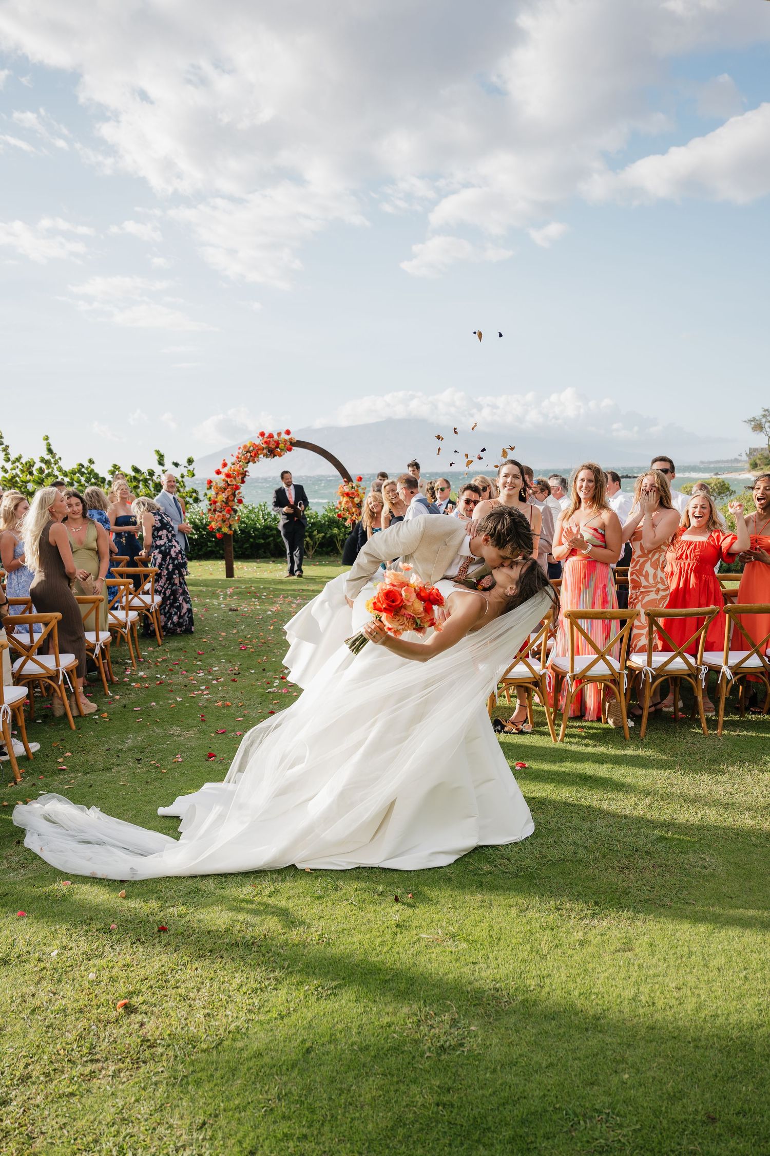 A bride's wedding dress flows dramatically in the wind during an outdoor ceremony with guests watching.