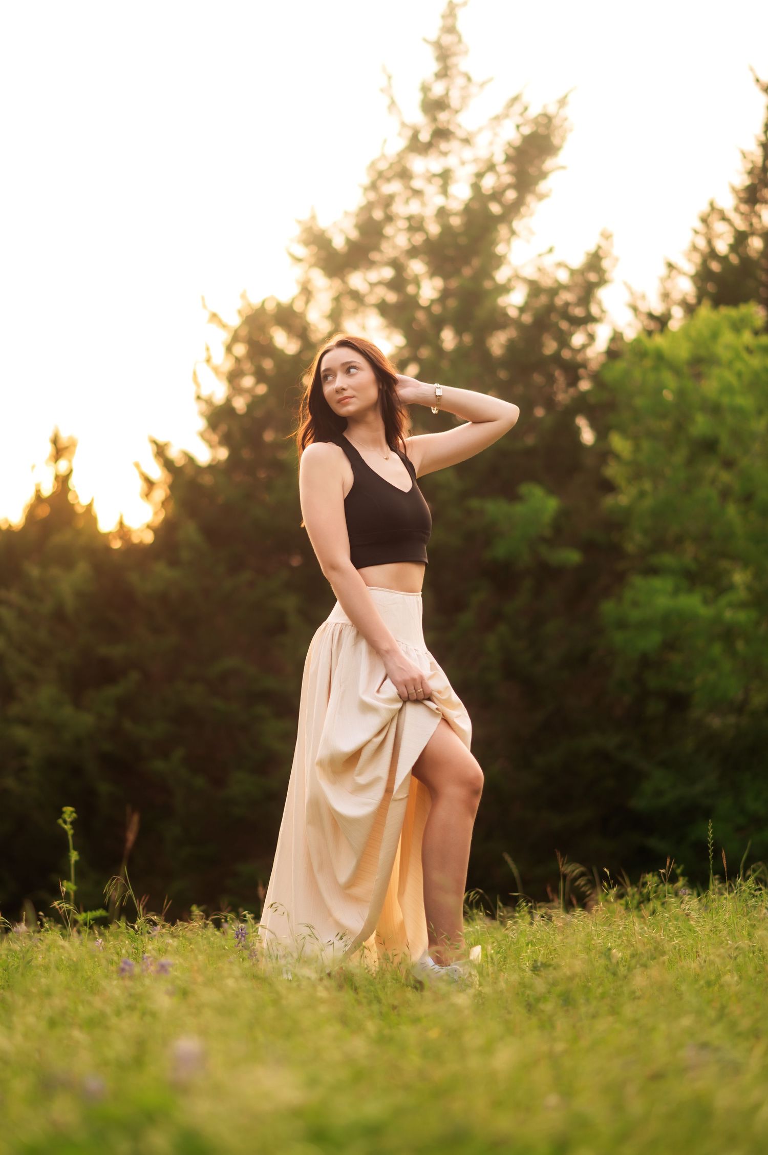 Young woman in black crop top and beige maxi skirt posing in a sunlit meadow with green trees at golden hour.