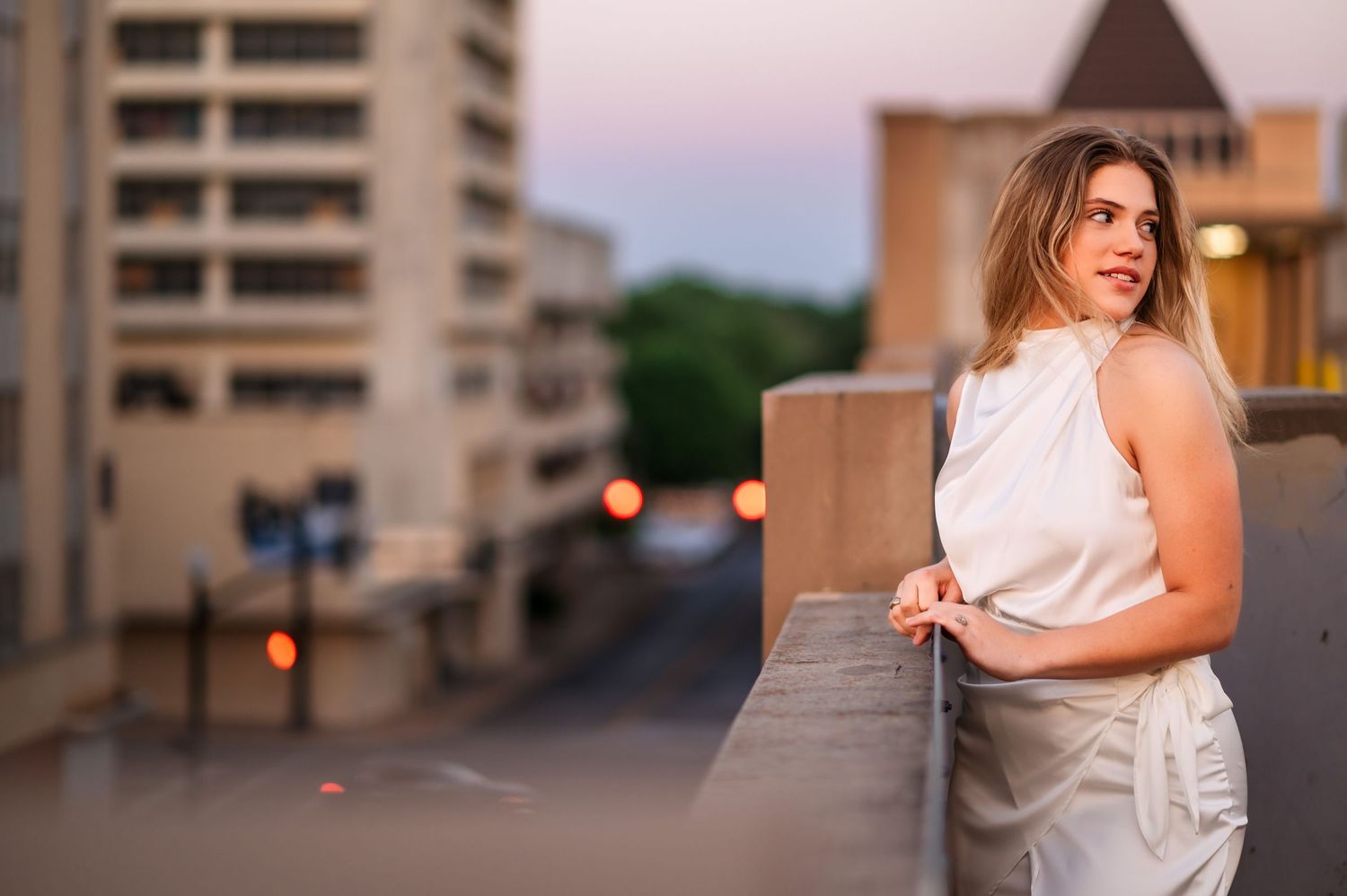 Woman in white sleeveless top leaning on rooftop railing at sunset in urban setting.