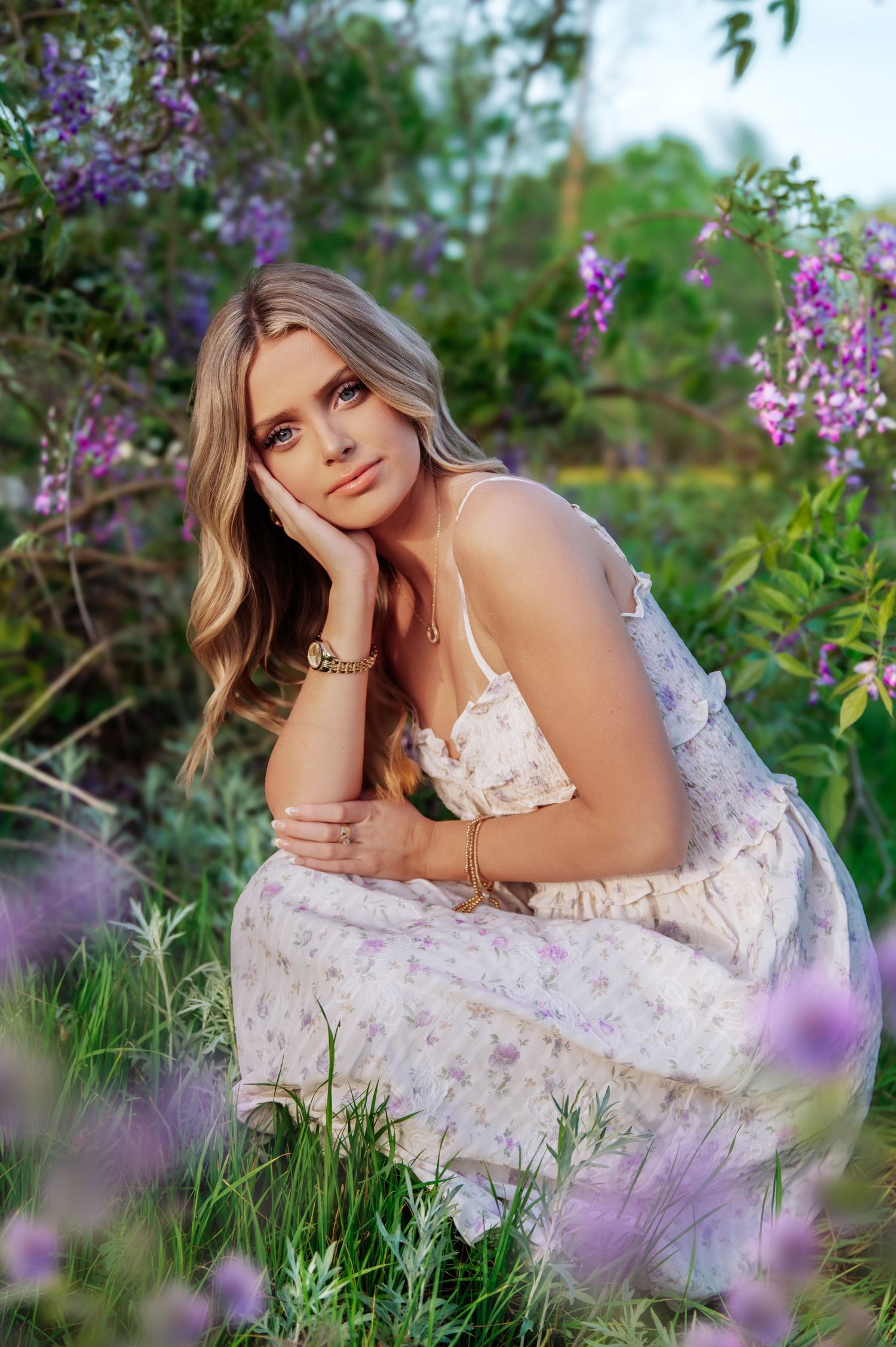 Young woman in floral dress sitting among purple wildflowers in a lush garden during golden hour.