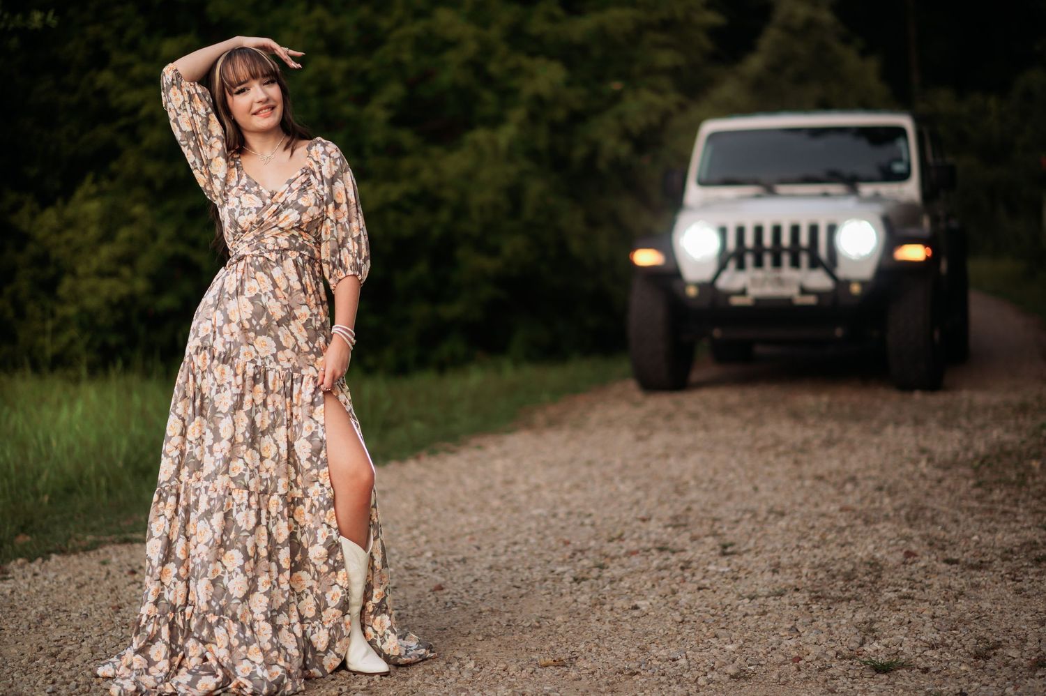 Woman in floral maxi dress and white boots poses on gravel path with Jeep Wrangler in background.