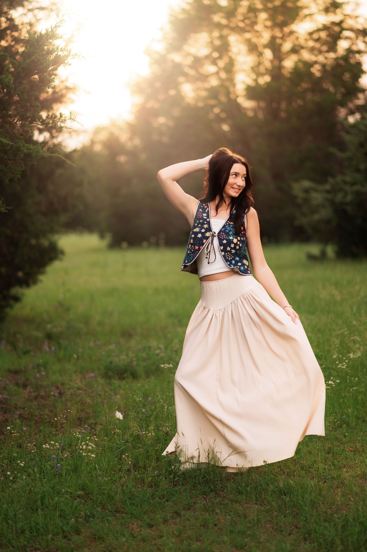 Woman in floral vest and flowing cream maxi skirt poses in sunlit green meadow at golden hour.