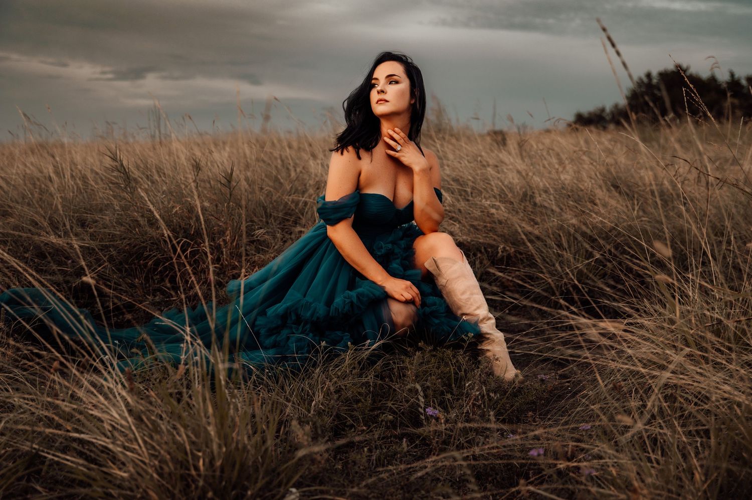 Woman in teal off-shoulder gown and white boots sitting gracefully in a golden grass field under dramatic cloudy skies.