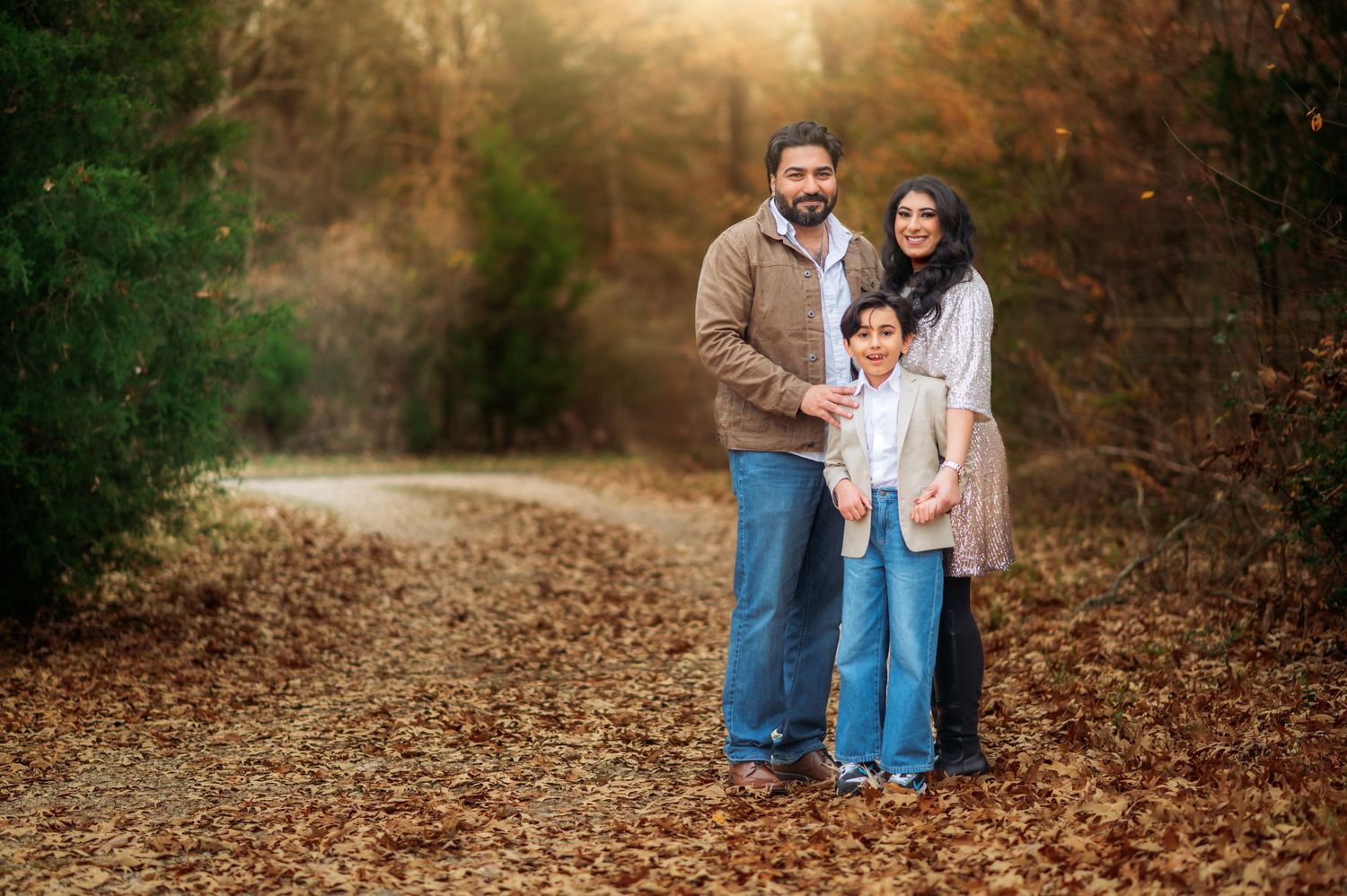 Family of three posing on a leaf-covered path surrounded by autumn trees in a warm outdoor portrait.