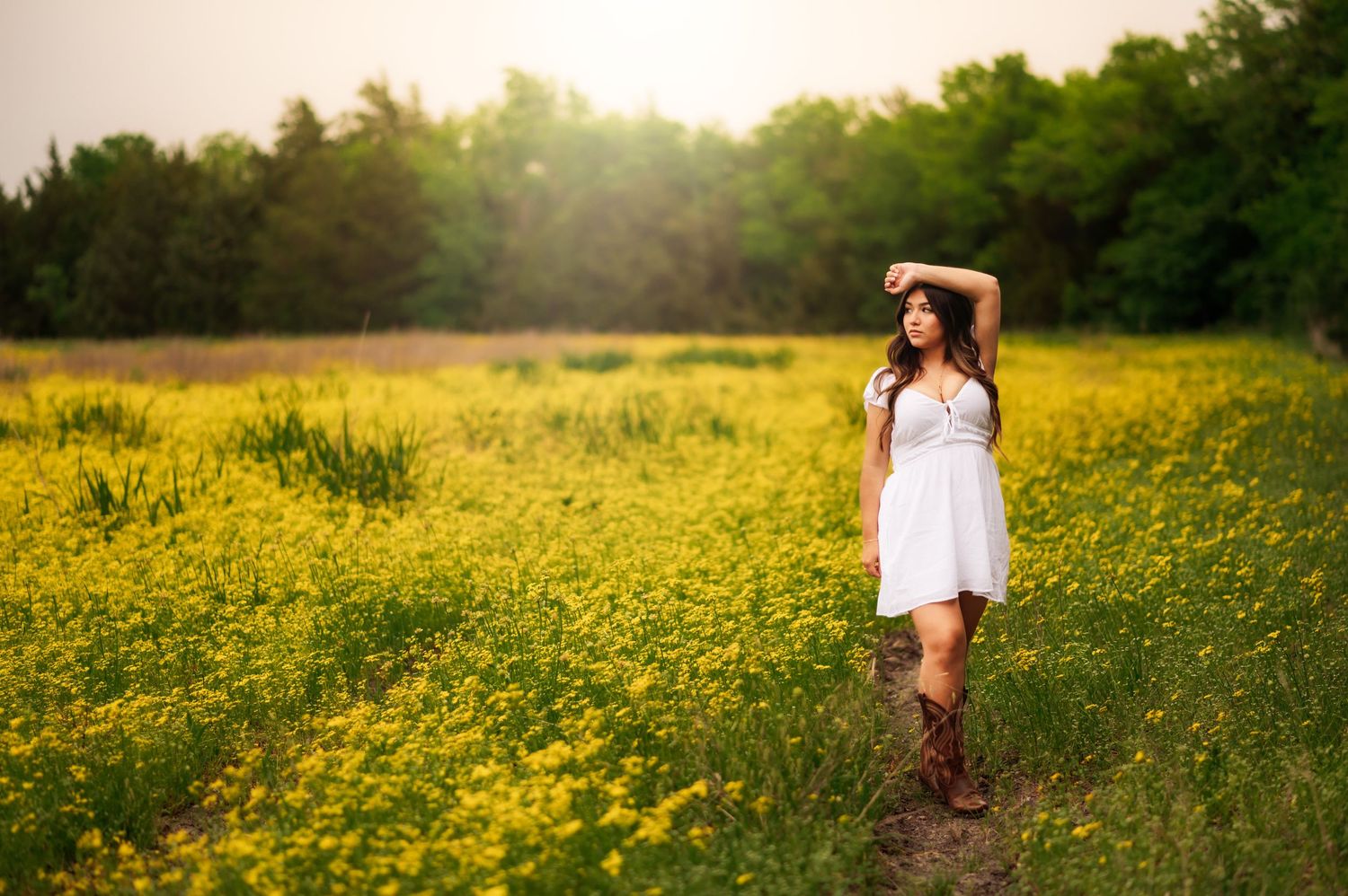Young woman in white dress and cowboy boots poses in a vibrant yellow wildflower field with lush green trees backdrop.