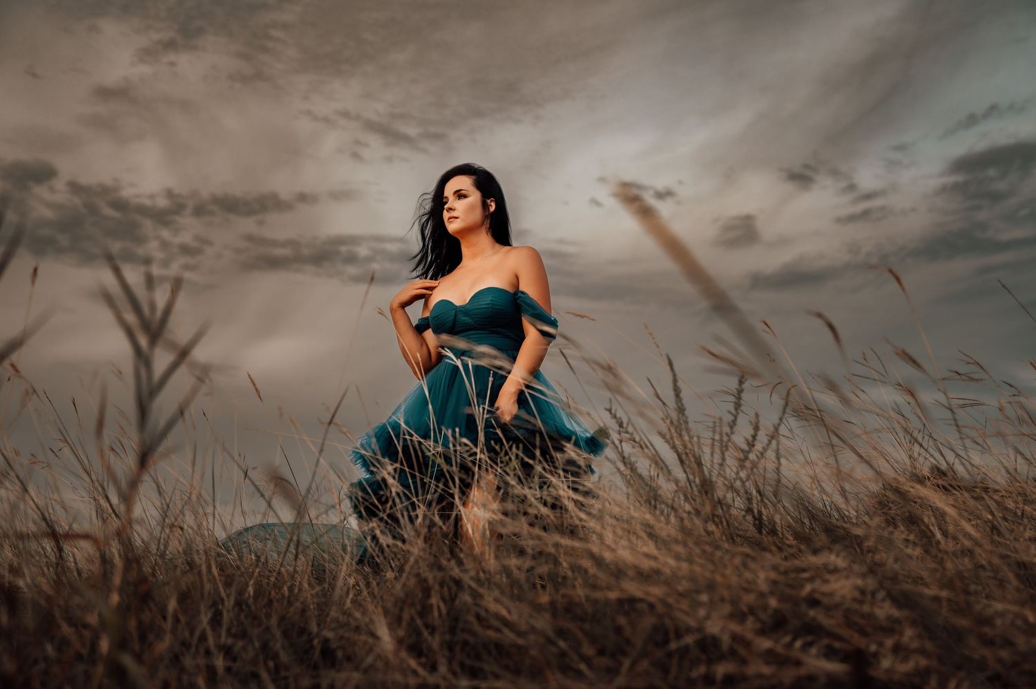 Woman in teal off-shoulder gown poses dramatically in golden grassland under stormy sky.