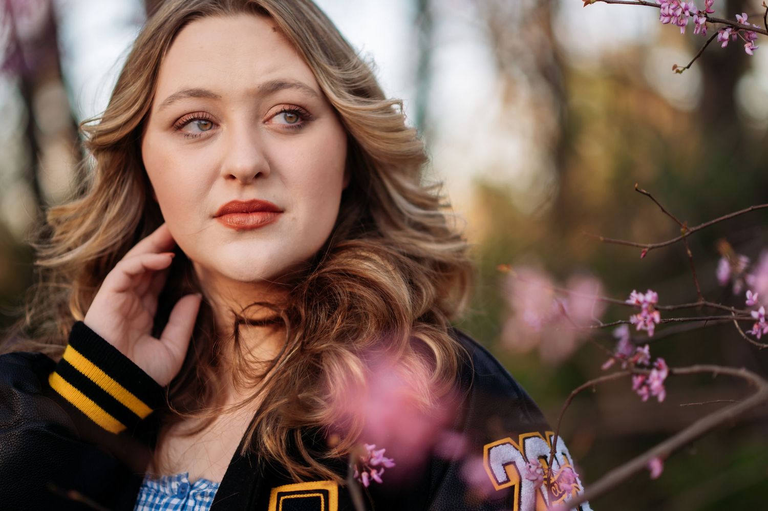 Young woman in varsity jacket poses near pink blossoms in soft natural light, spring outdoor portrait photography.