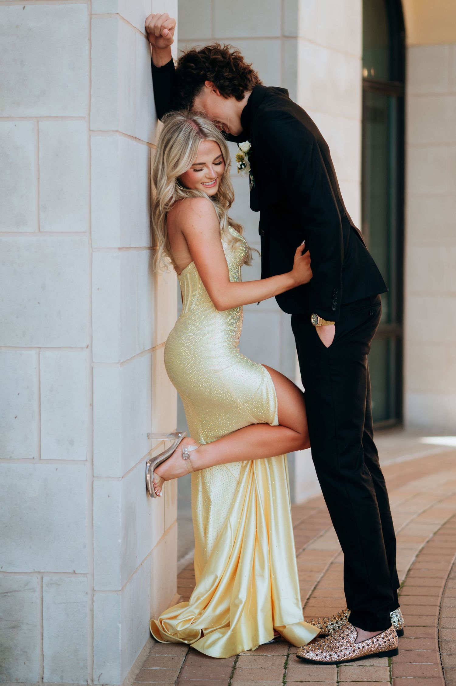Couple in formal wear posing against a wall, woman in yellow satin gown, man in black suit kissing her cheek.