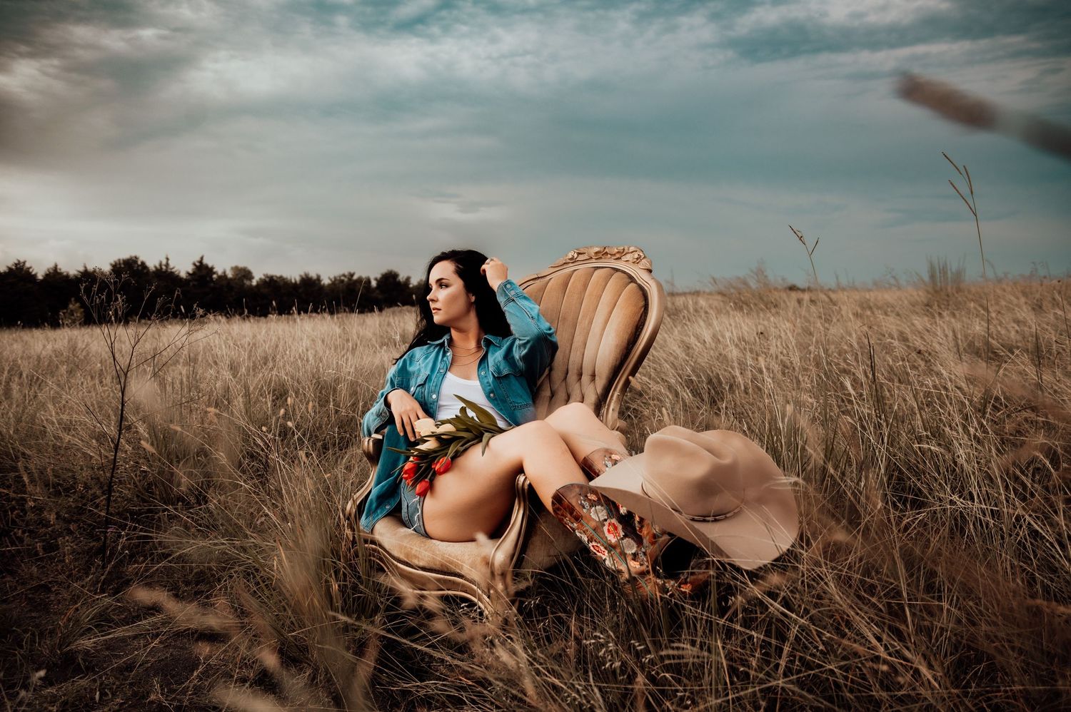 Woman in denim jacket sitting in ornate chair in golden field holding book, cowboy hat nearby, dramatic cloudy sky.