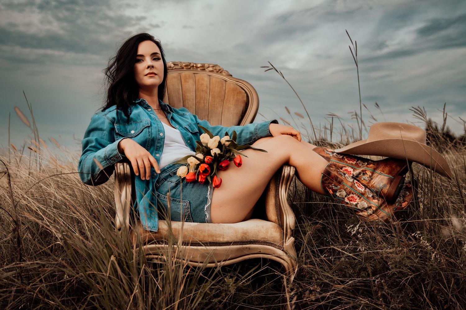 Woman in denim jacket reclining in ornate chair surrounded by tall grass in open field.