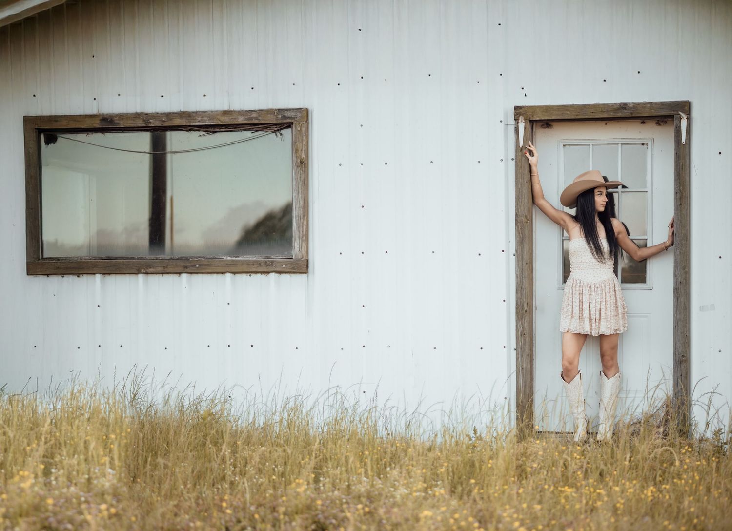 A woman in a cowboy hat and white dress poses in a doorway of a rustic white barn amid golden grass.