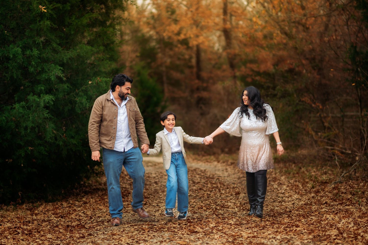 Family of three walking hand-in-hand through autumn forest path covered in fallen leaves.