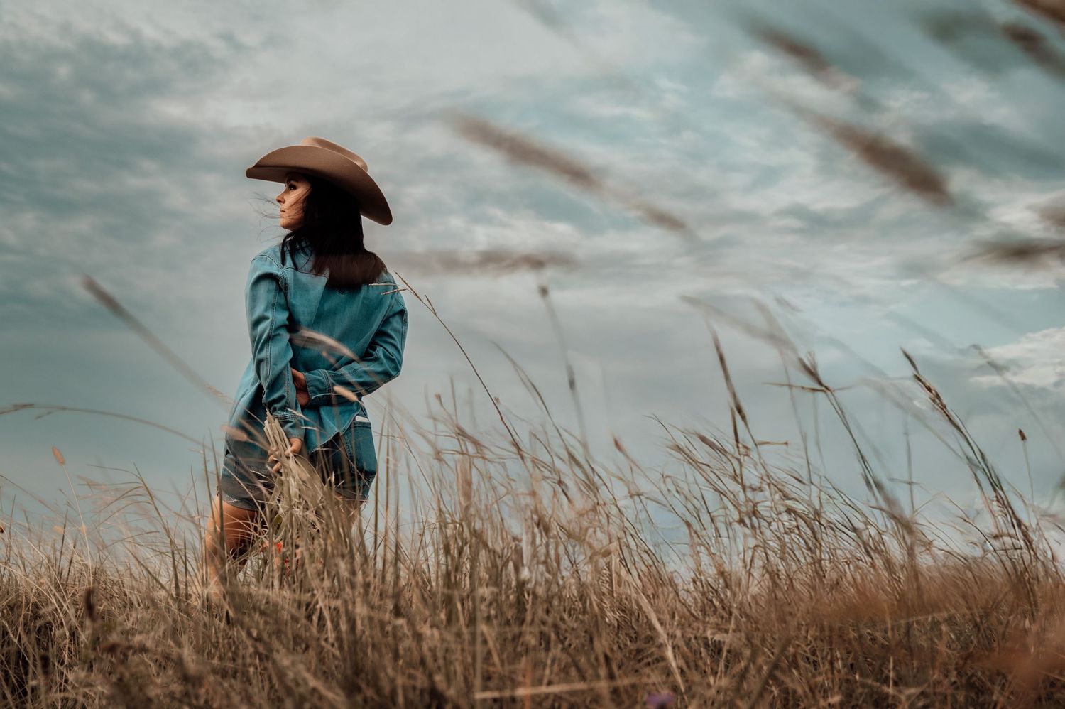 Woman in teal jacket and cowboy hat standing in golden grass field under cloudy sky.