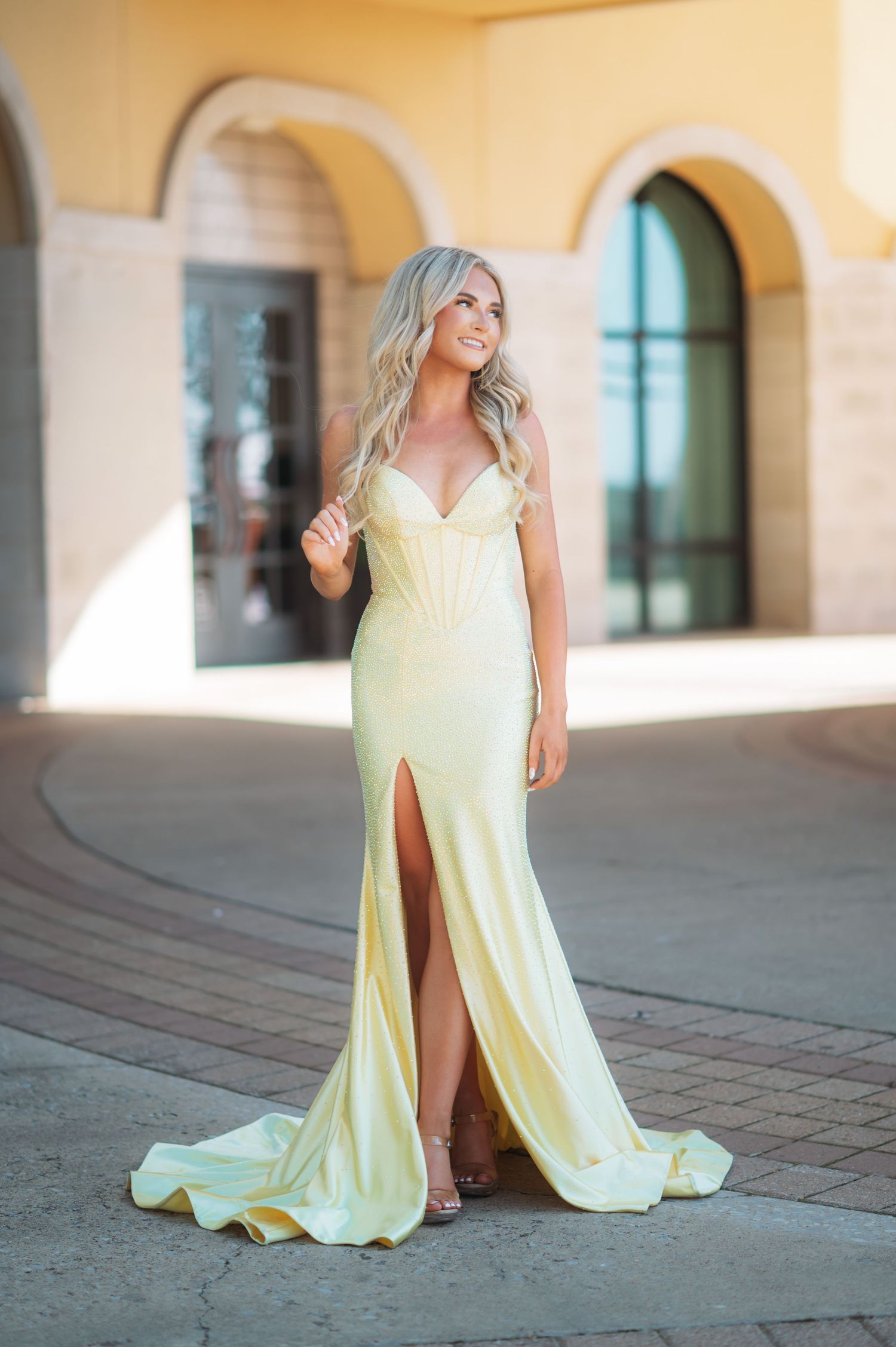 Woman in yellow satin strapless gown with thigh slit posing outdoors near arched building.
