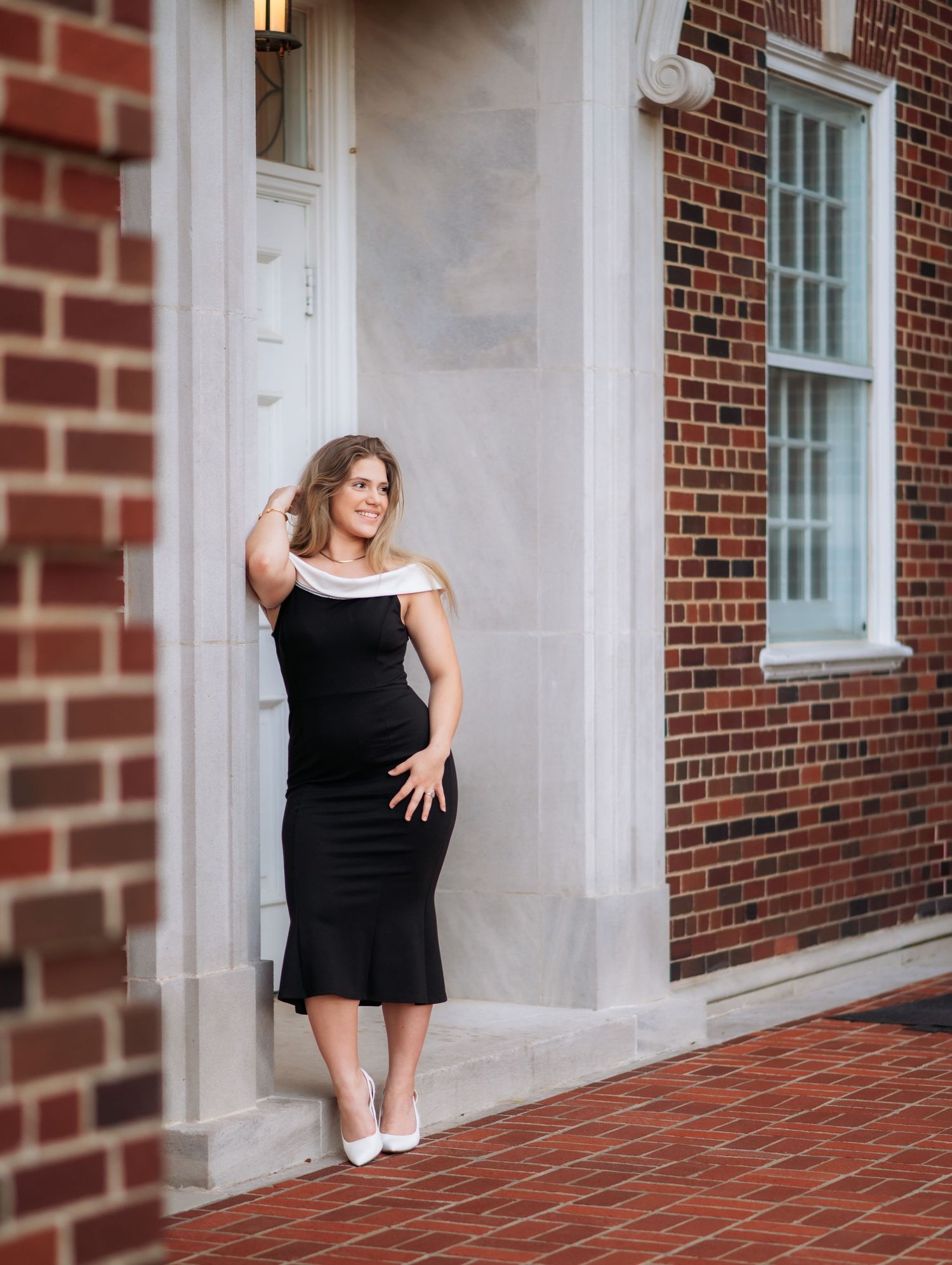 Woman in black midi dress with white heels posing by white door of brick building.