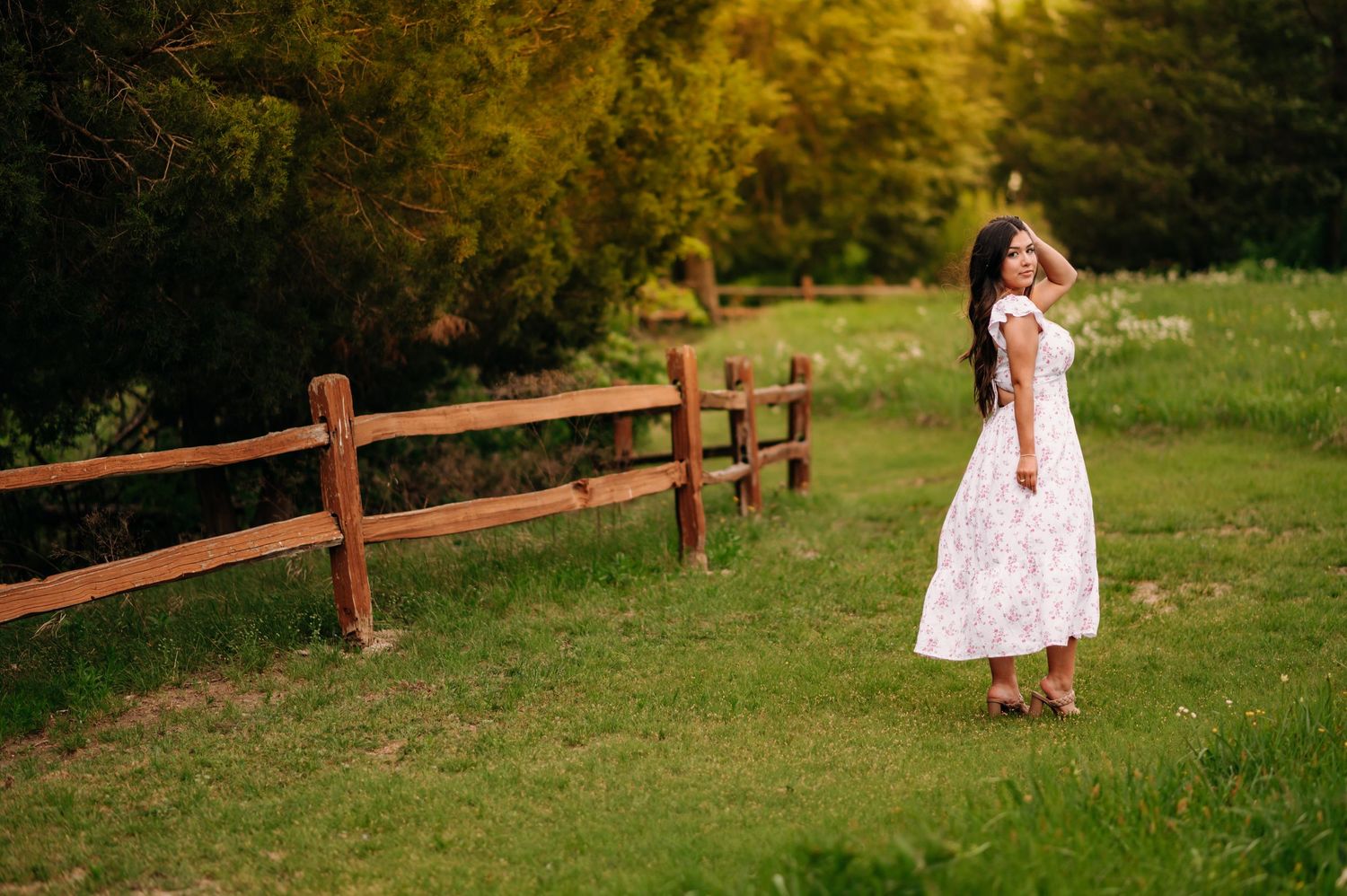 A young girl in a white floral dress stands by a wooden fence in a lush green meadow at golden hour.