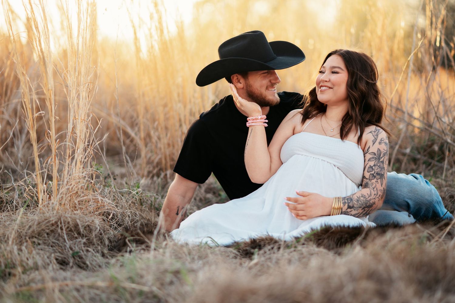Couple sitting in golden field at sunset, man in black cowboy hat, pregnant woman in white dress with tattoos.