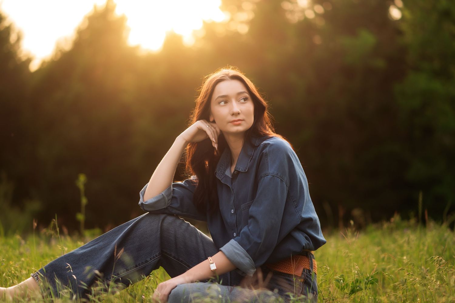 Young woman in denim outfit sitting in sunlit meadow at golden hour, gazing thoughtfully into the distance.