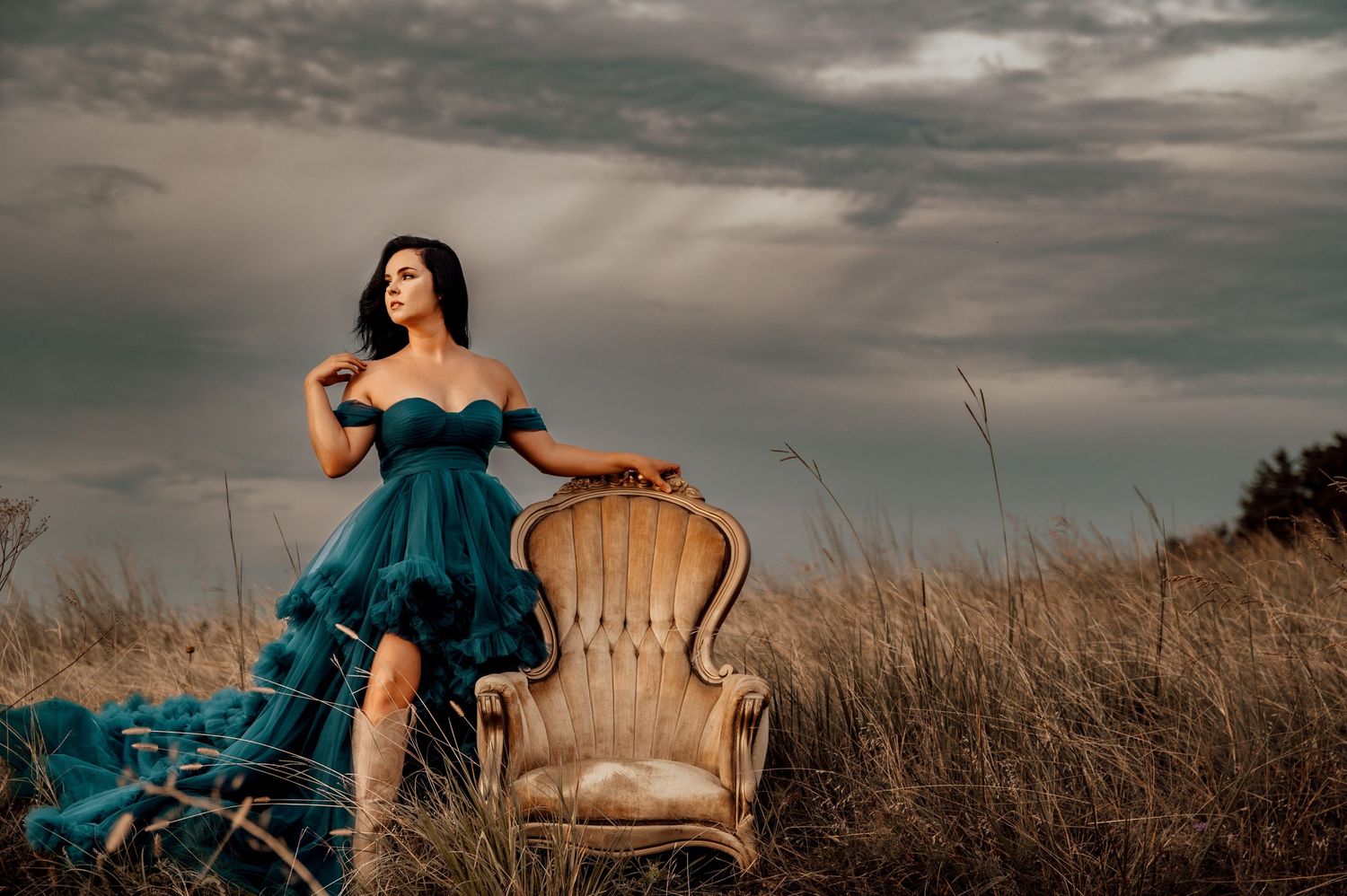 Woman in teal off-shoulder gown poses beside vintage armchair in golden field under dramatic cloudy sky.