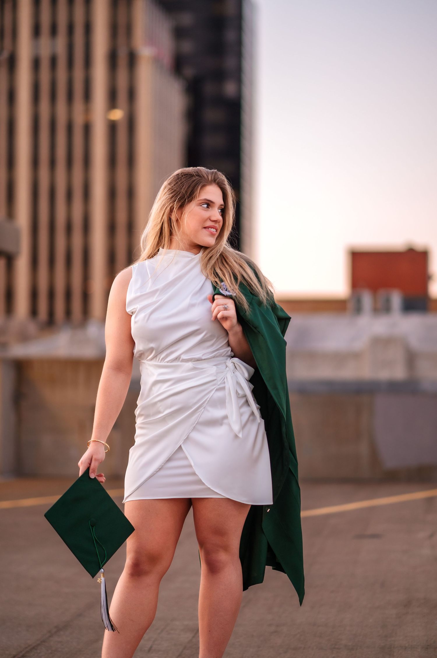Young woman in white dress holding green graduation cap and gown on rooftop at sunset.