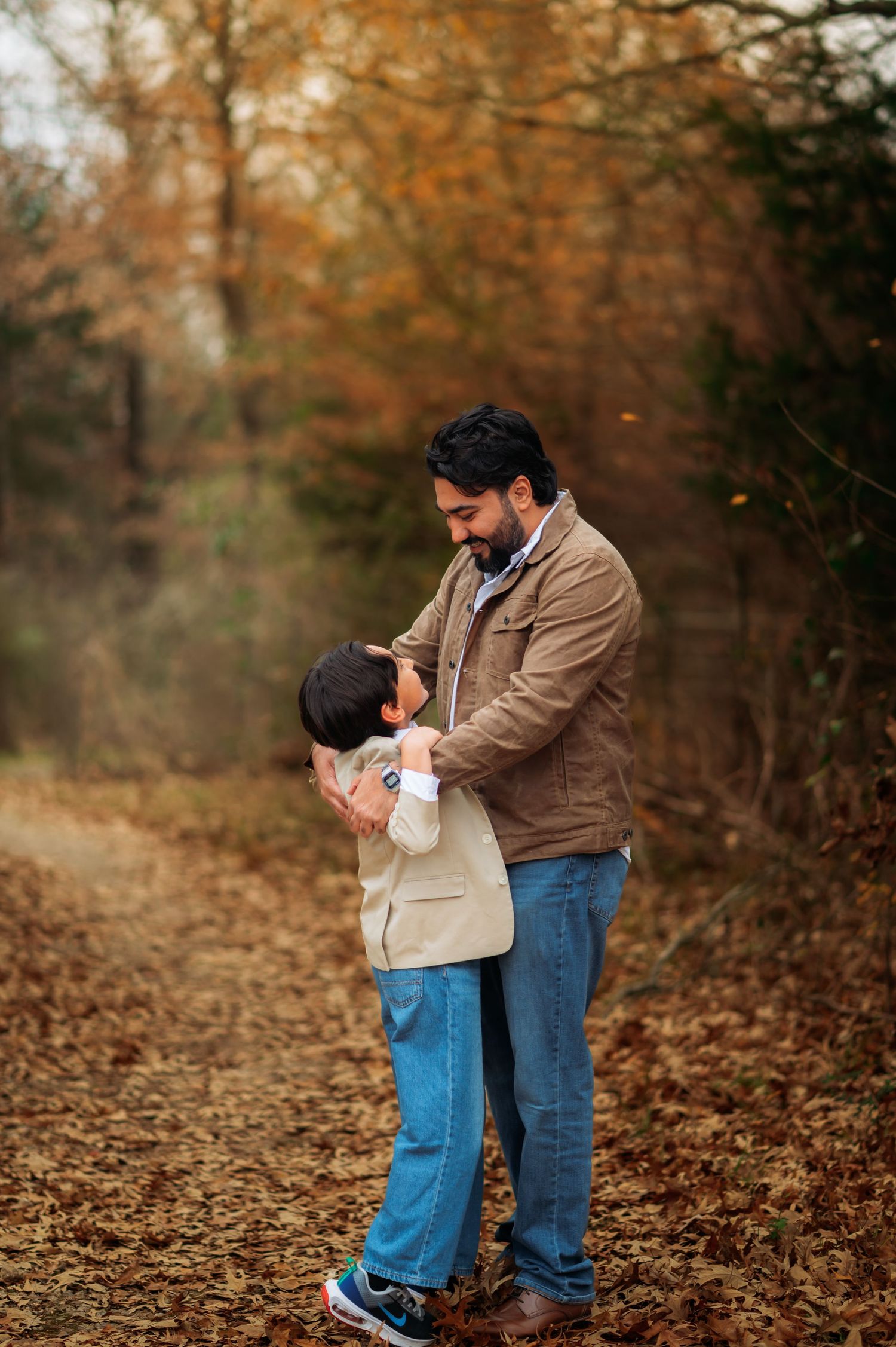 A father and child share a warm embrace on a leaf-covered path surrounded by autumn foliage.