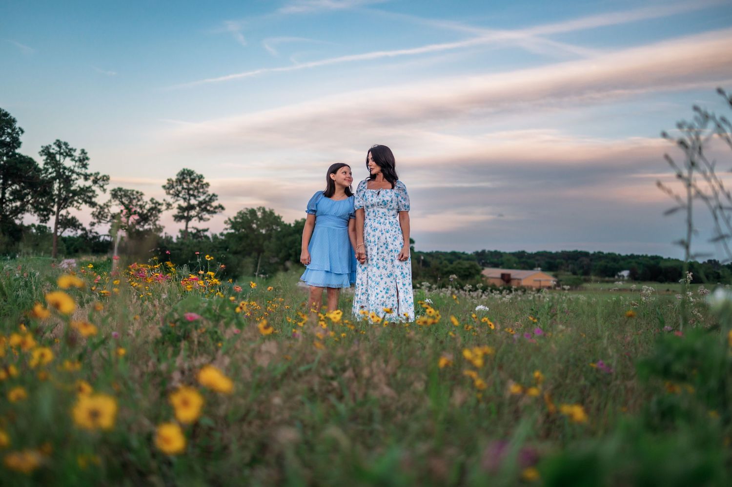 Two women in summer dresses stand in a wildflower meadow at sunset, sharing a warm moment together.