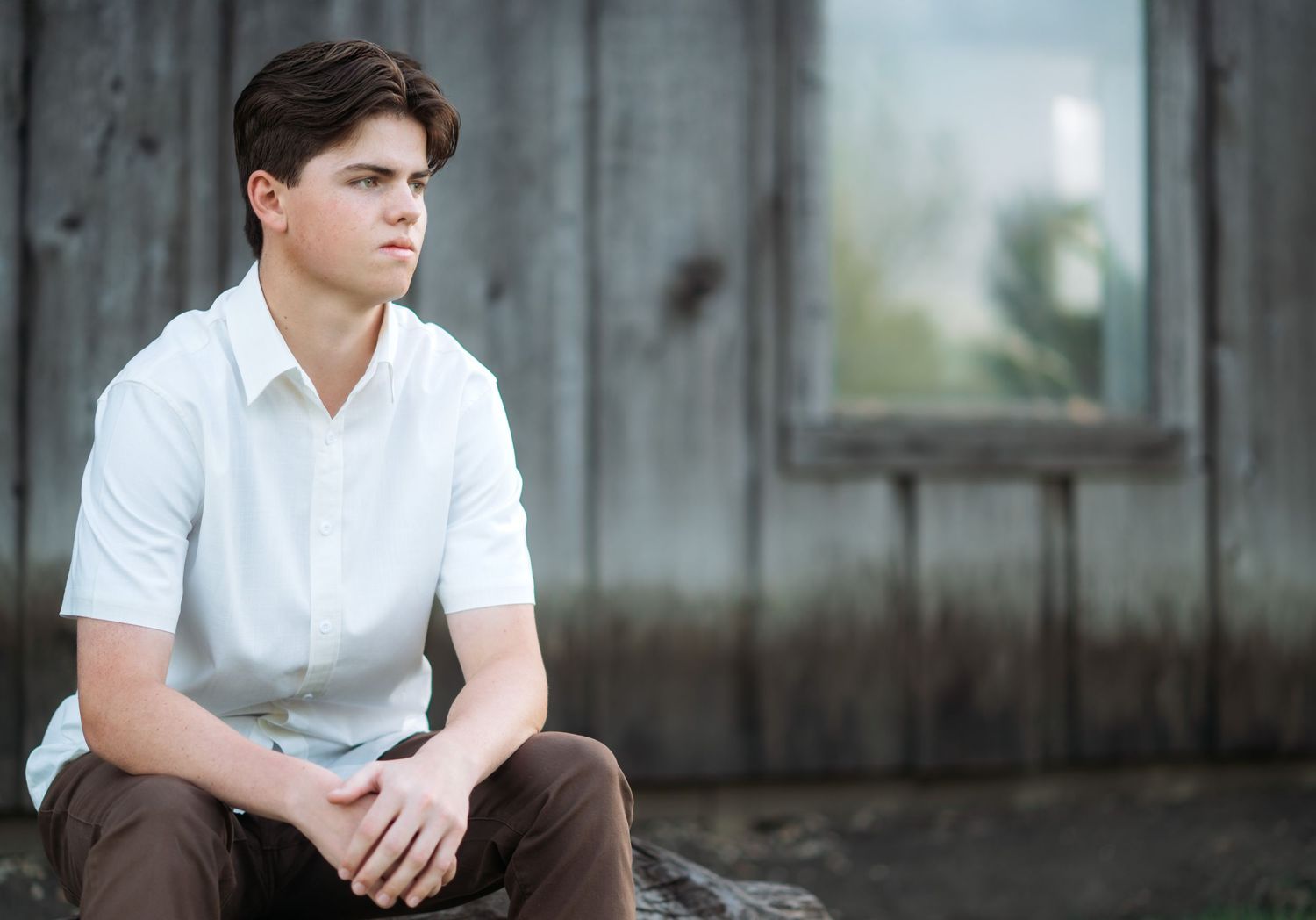 Young man in white shirt and brown pants sitting outdoors near rustic wooden wall, looking thoughtfully to the side.