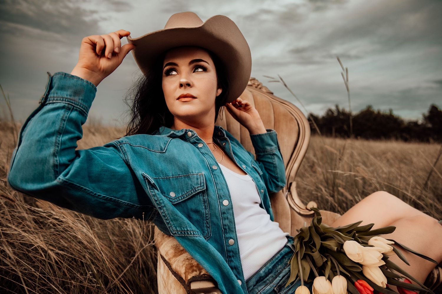 A young woman in a denim jacket and cowboy hat sits in a field chair holding white tulips under a dramatic cloudy sky.