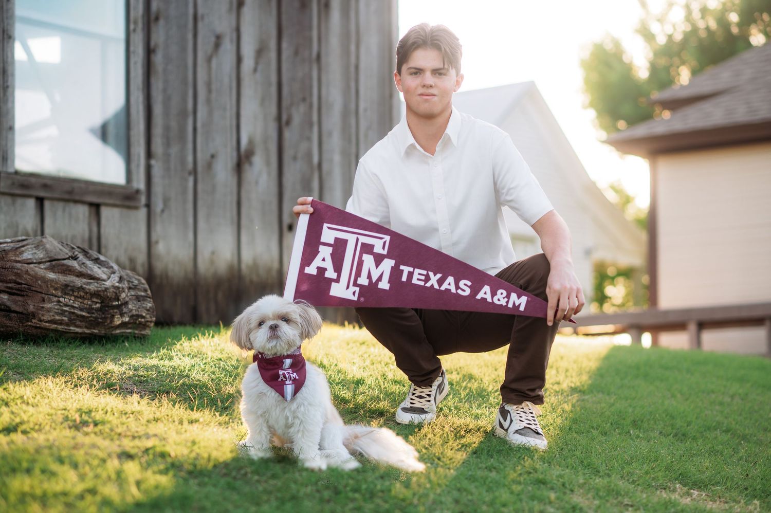 Young man holding Texas A&M pennant poses with small white dog on grass near rustic barn.